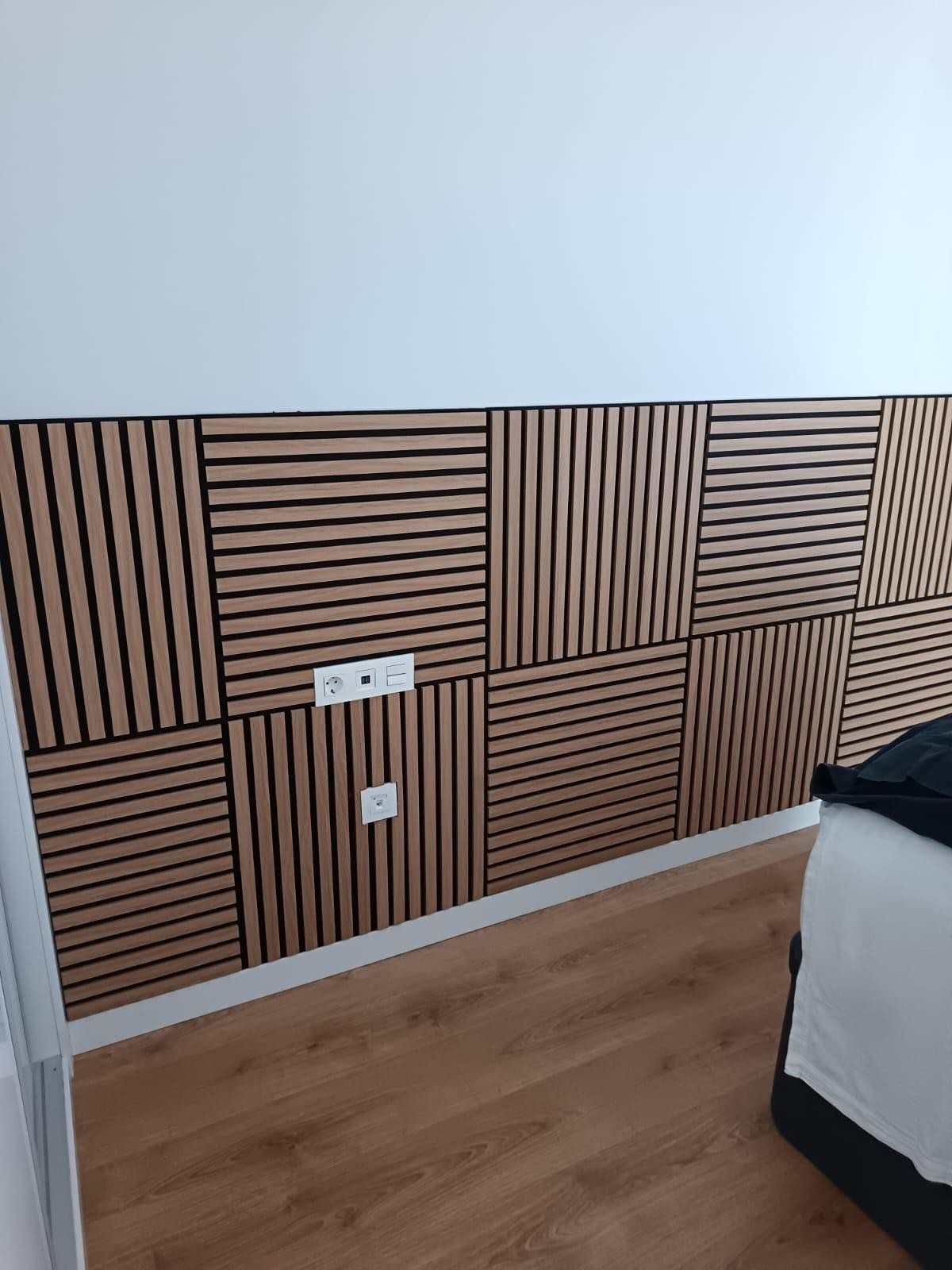 Pared de dormitorio revestida de madera con listones geométricos, enchufes y parte de una cama sobre un suelo de madera.