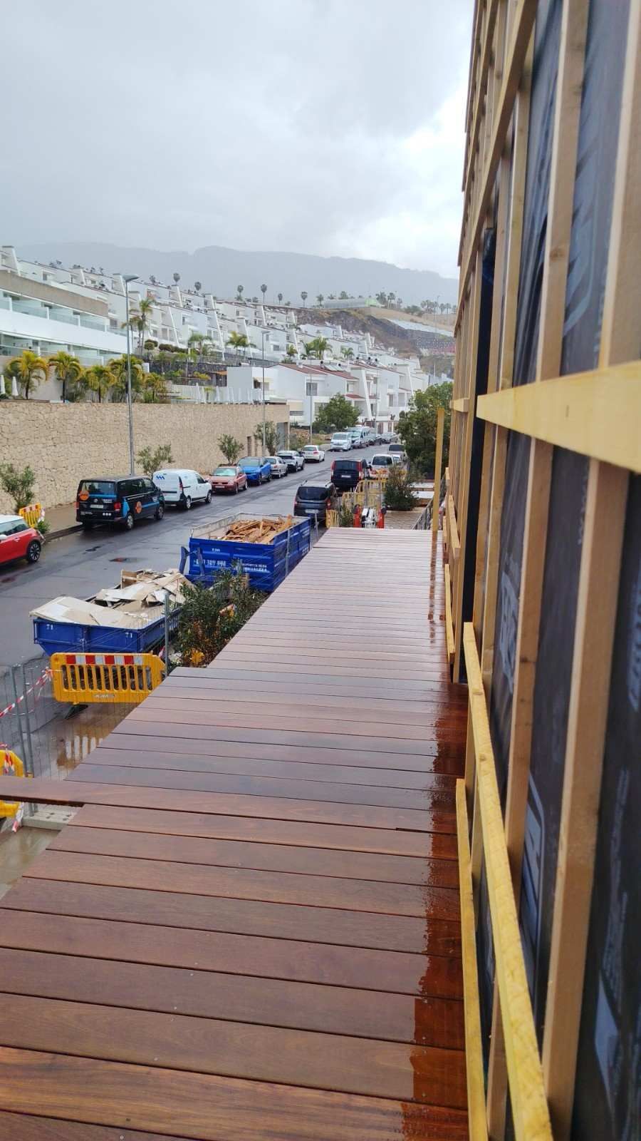 Pasarela de madera mojada junto a coches aparcados en una calle de ladera bajo un cielo nublado.