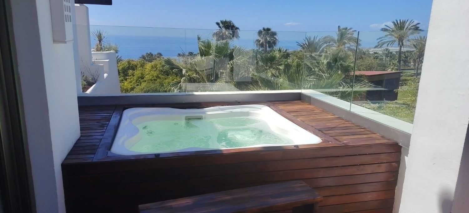 Pequeña bañera de hidromasaje de madera en un balcón con vistas al océano y a las palmeras.