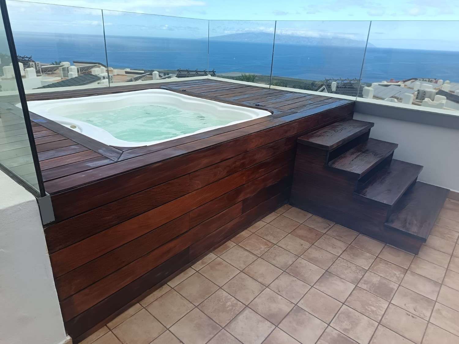 Terraza en la azotea con jacuzzi de madera, escalones oscuros y vistas al océano más allá de un patio embaldosado.