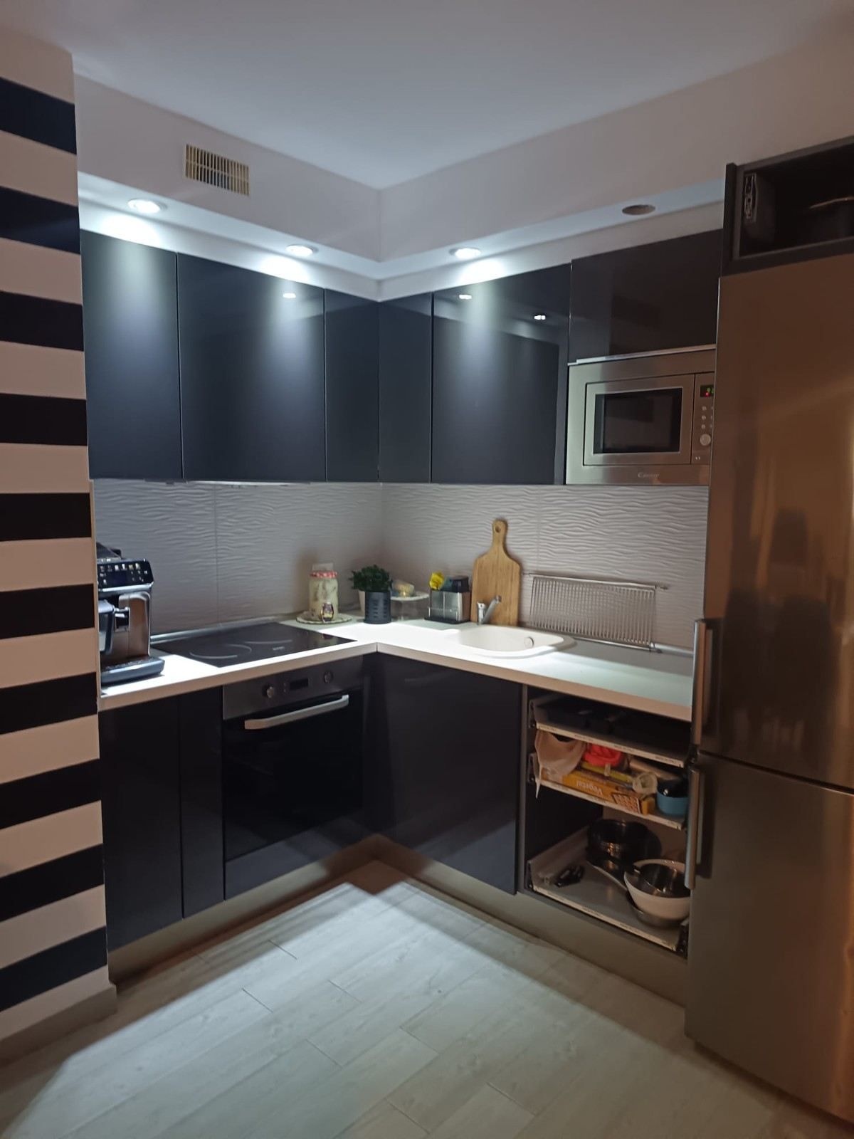 Cocina moderna con armarios oscuros, encimeras blancas, nevera de acero inoxidable e iluminación bajo los armarios.