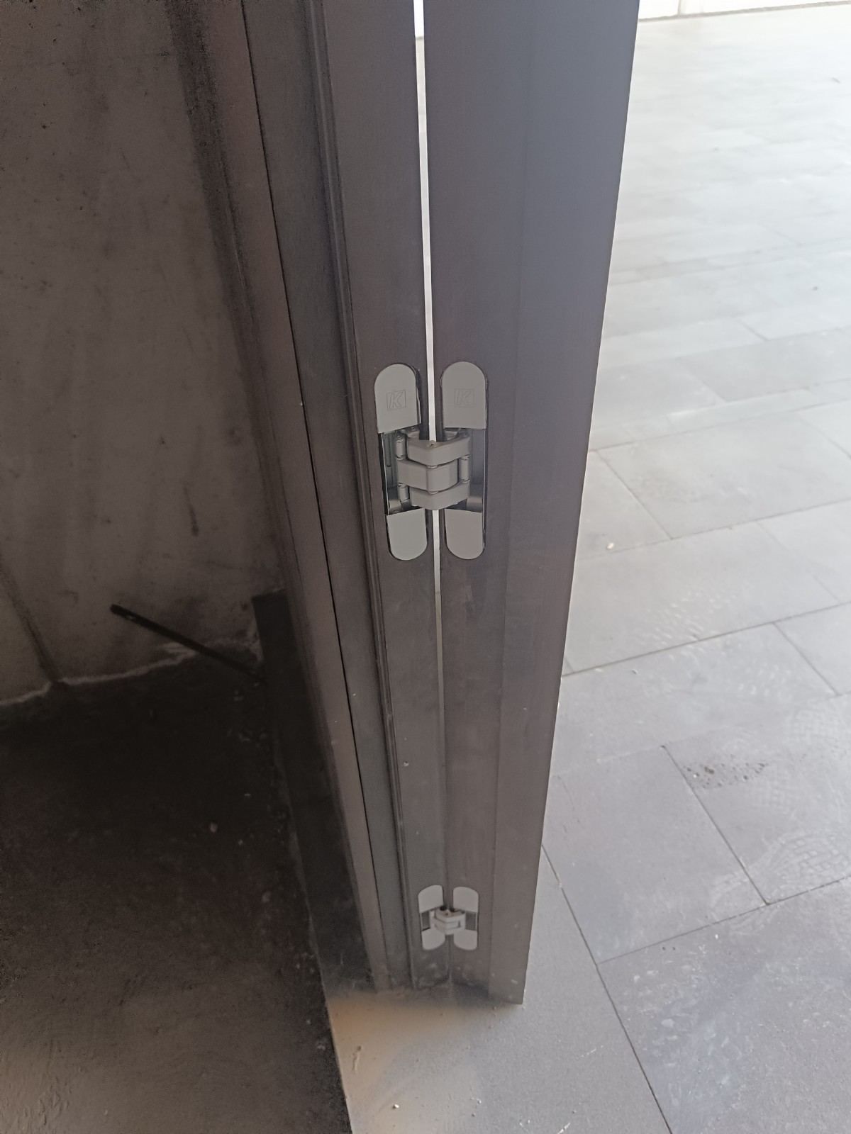 Primer plano de una bisagra de puerta gris en una puerta parcialmente abierta con suelo de baldosas.