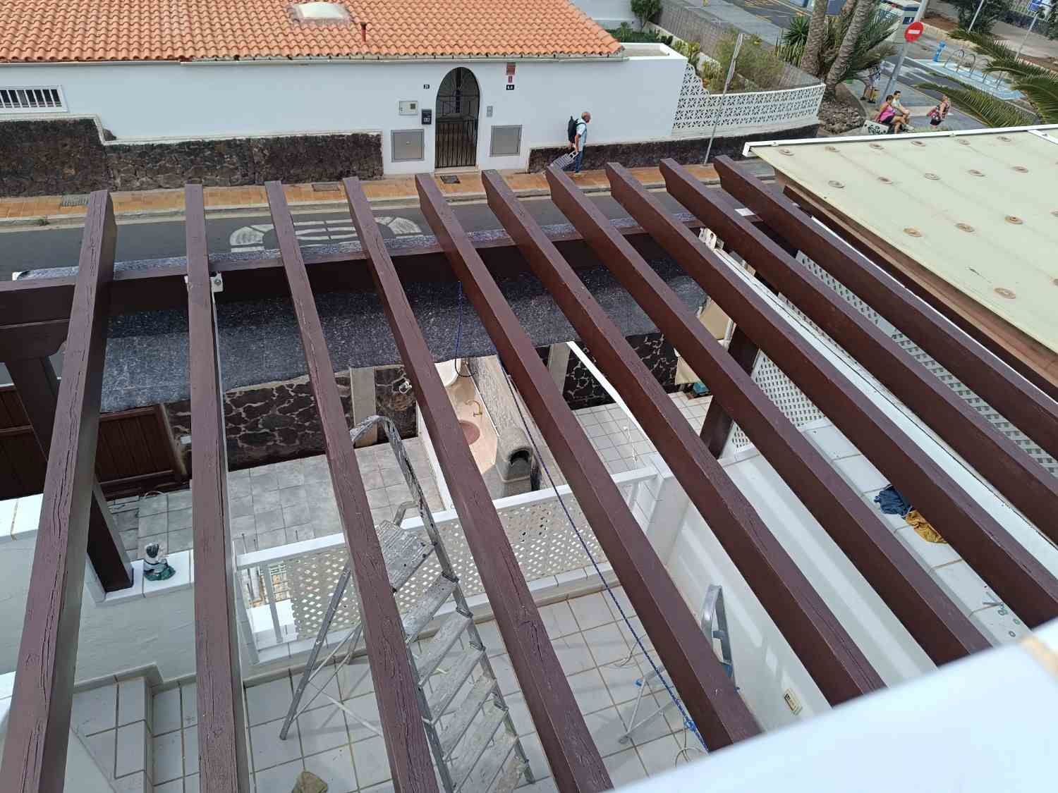 Vigas de madera en una pérgola con vistas a un patio embaldosado y a un edificio blanco cercano con tejado de terracota.