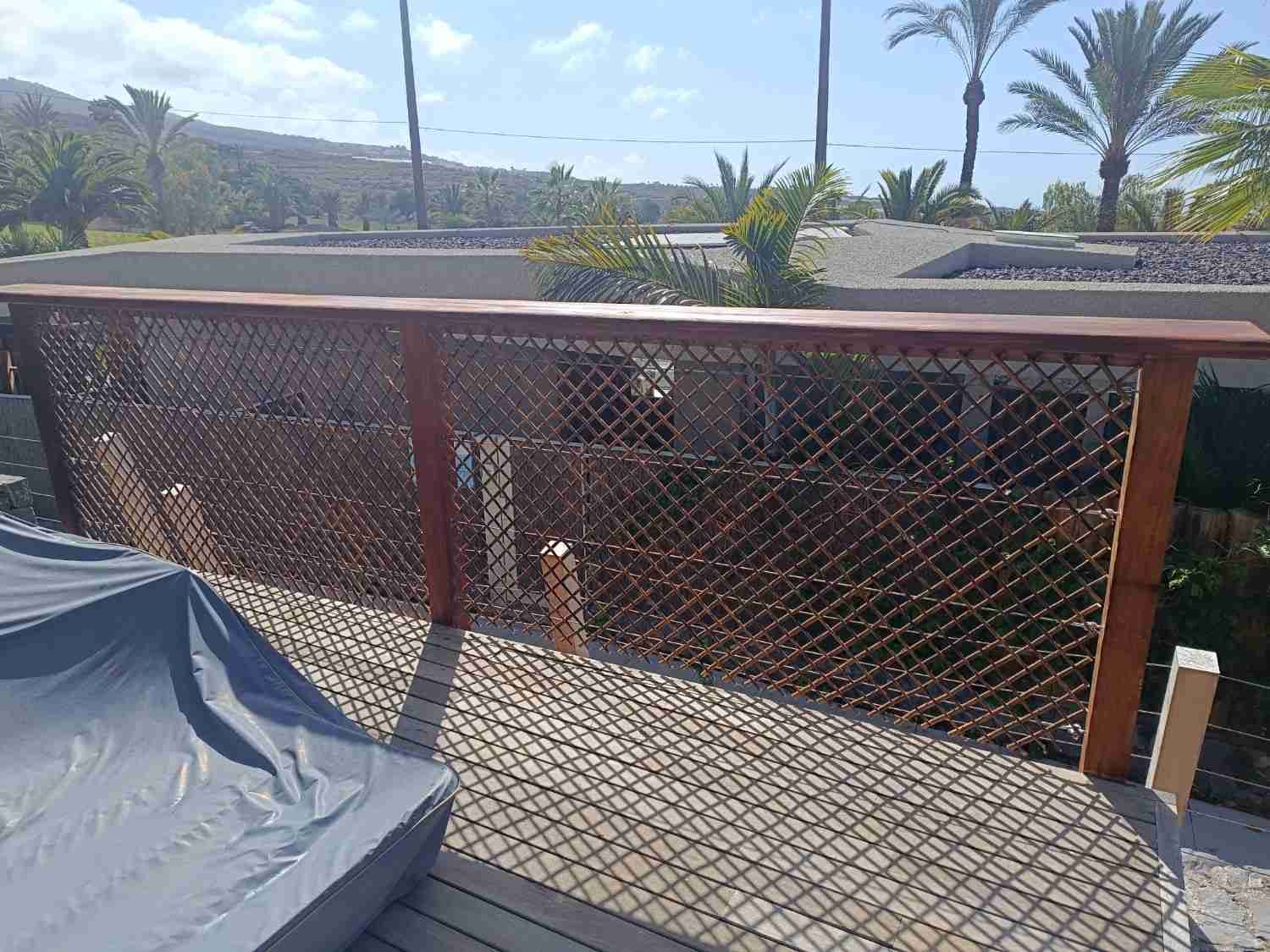 Barandilla de balcón de madera con malla metálica con vistas a palmeras bañadas por la luz del sol.