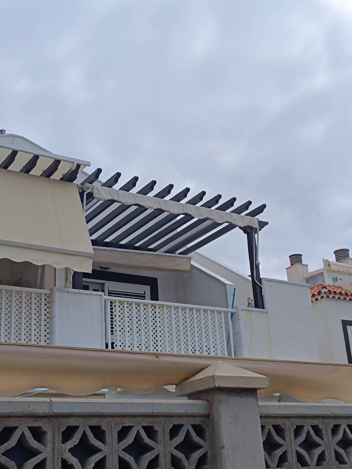Balcón con barandilla blanca y pérgola a rayas bajo un cielo nublado.