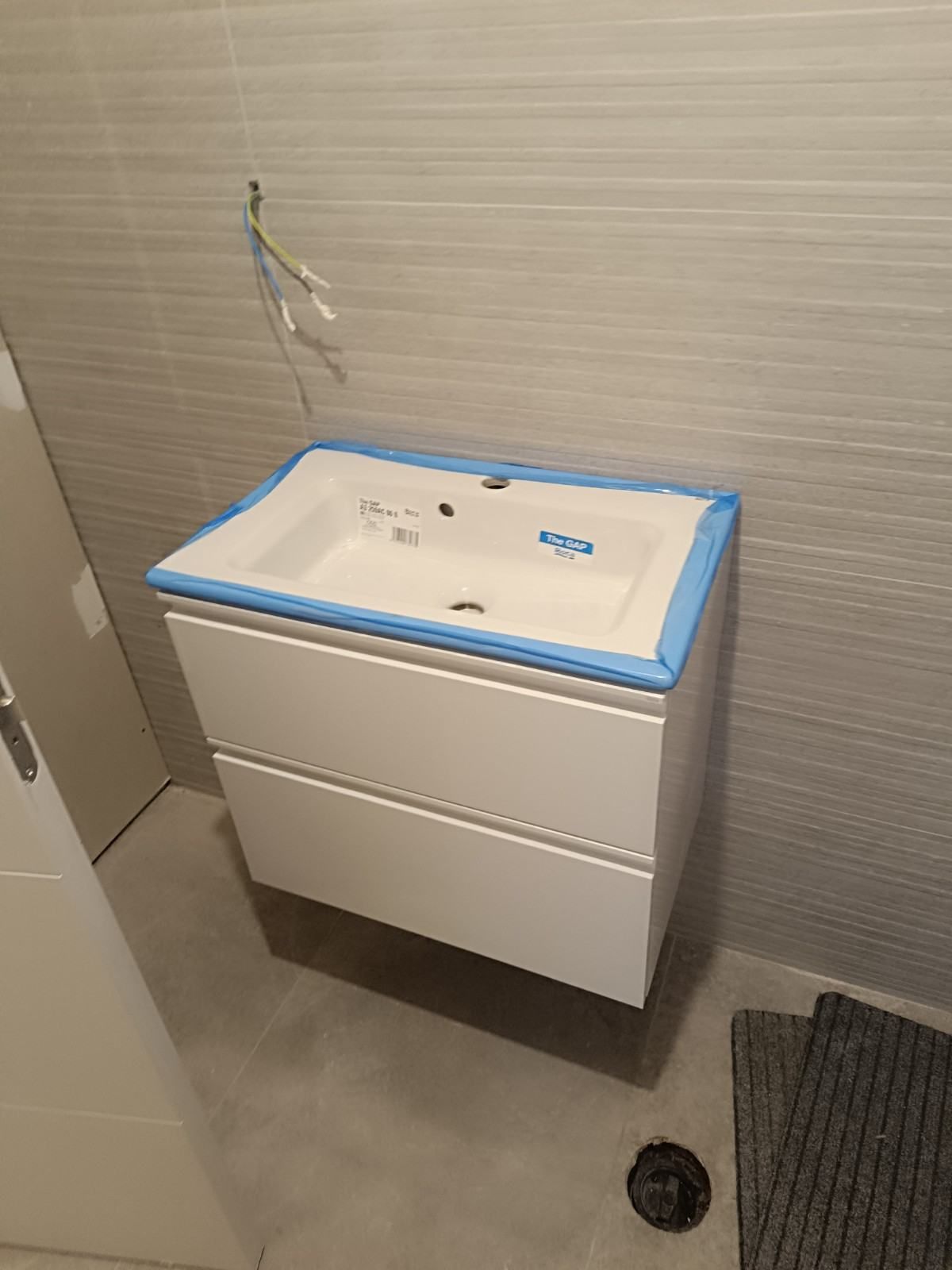 Mueble de baño pequeño de color blanco con lavabo y tuberías a la vista, colocado sobre una pared alicatada, con cinta adhesiva azul en la parte superior.