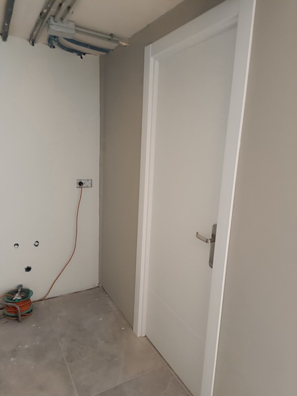 Habitación vacía en obras con techo sin terminar, puerta blanca y cableado expuesto en el suelo de hormigón.