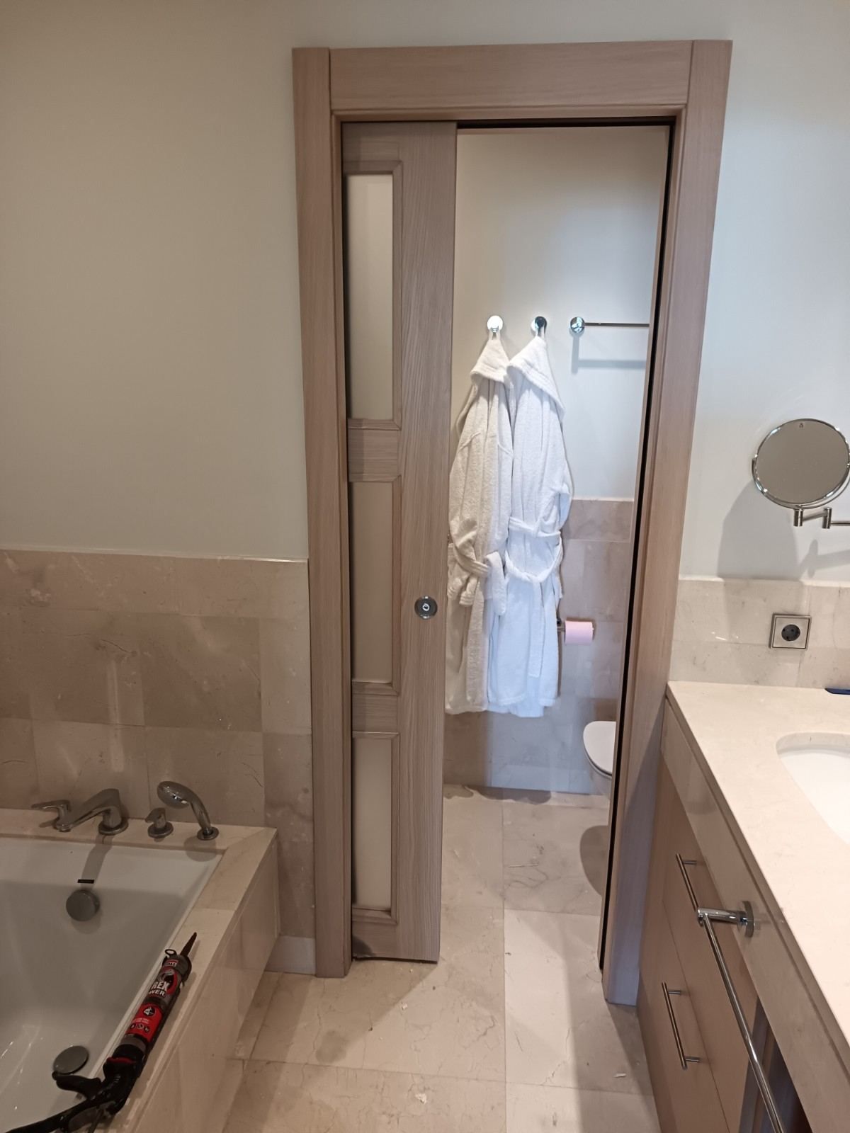 Entrada del baño con una bata blanca colgada de un gancho, espejo, lavabo y bañera a la vista.