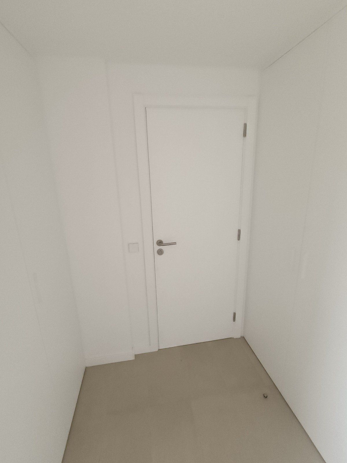 Pequeño pasillo blanco con una puerta blanca cerrada y suelo beige.