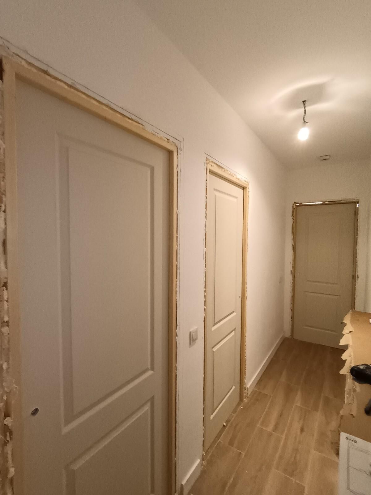 Pasillo estrecho con puertas blancas, paredes beige y una lámpara de techo, con vista hacia una puerta abierta.