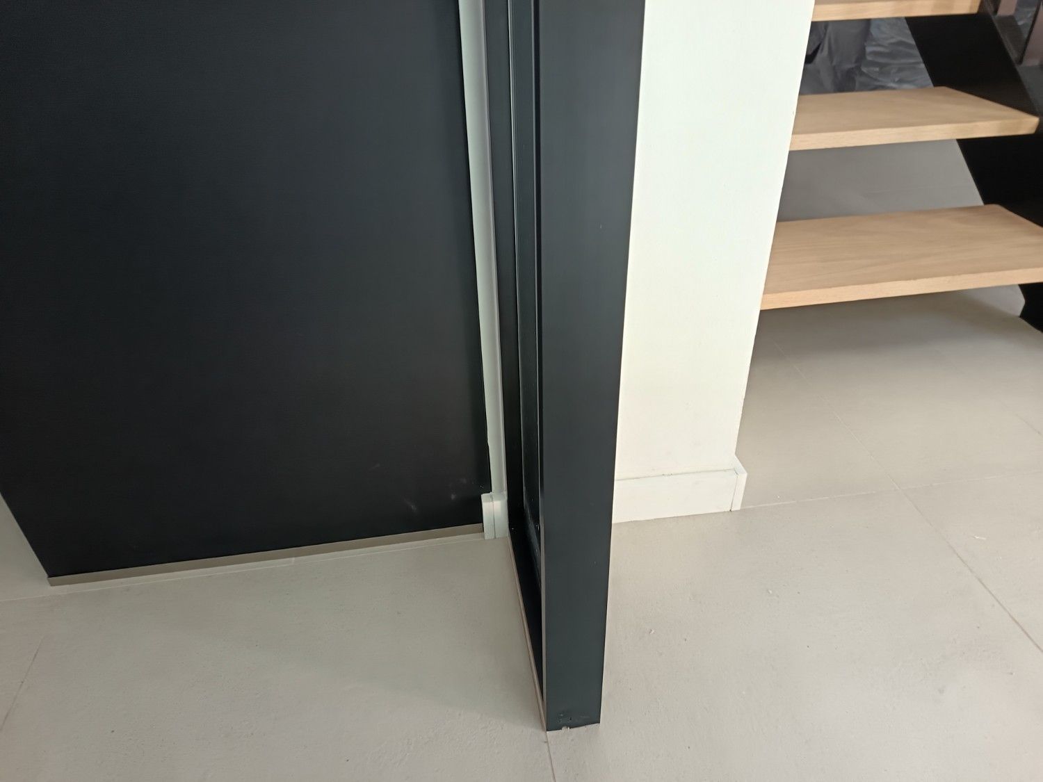 Escalera moderna con escalones de madera clara, panel de pared oscuro y suelo de baldosas claras junto a una pared blanca.
