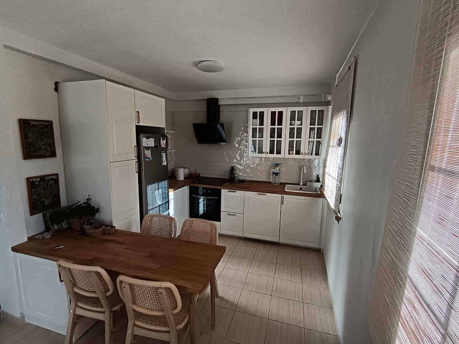 Cocina pequeña con mesa de madera, armarios blancos, suelo de baldosas y persianas que dejan pasar la luz.