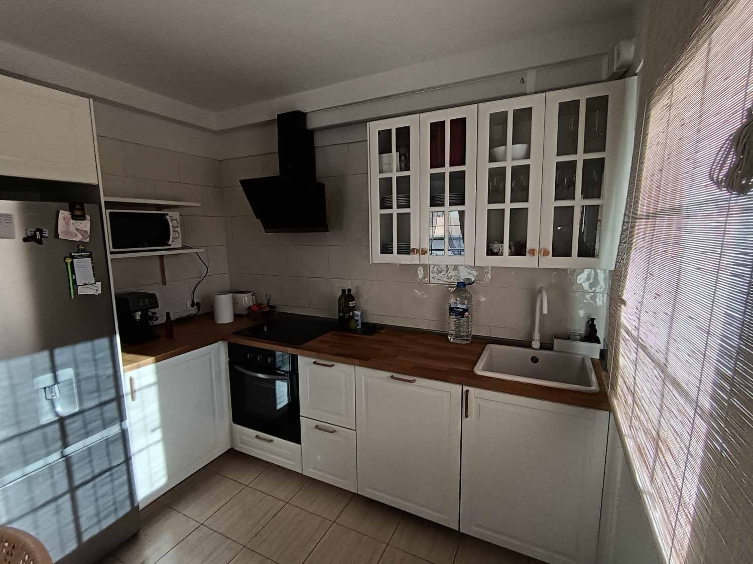 Pequeña cocina blanca con encimeras y armarios de madera, suelo de baldosas y un armario con ventana encima del fregadero.