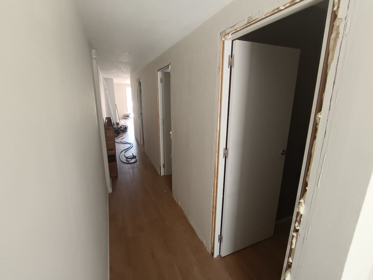 Pasillo estrecho con paredes blancas, suelo de madera y puertas abiertas al final.