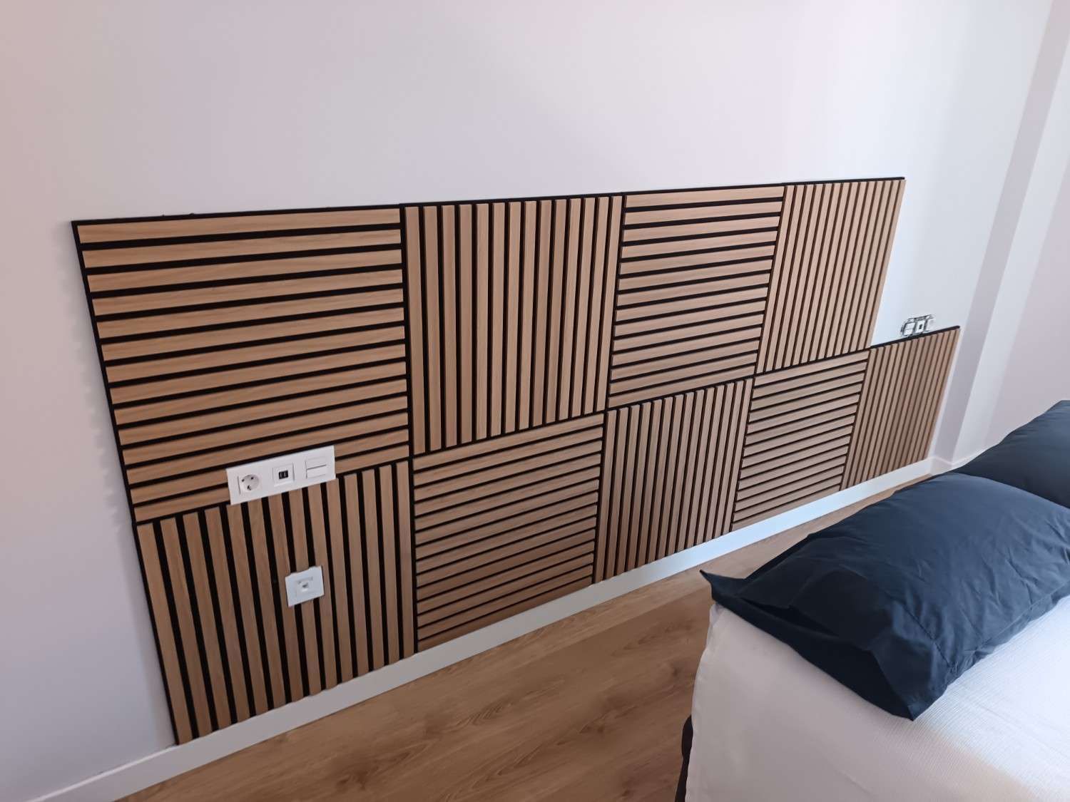 Panel decorativo de listones de madera sobre una cama en un dormitorio minimalista, con un enchufe blanco y una almohada oscura cerca.