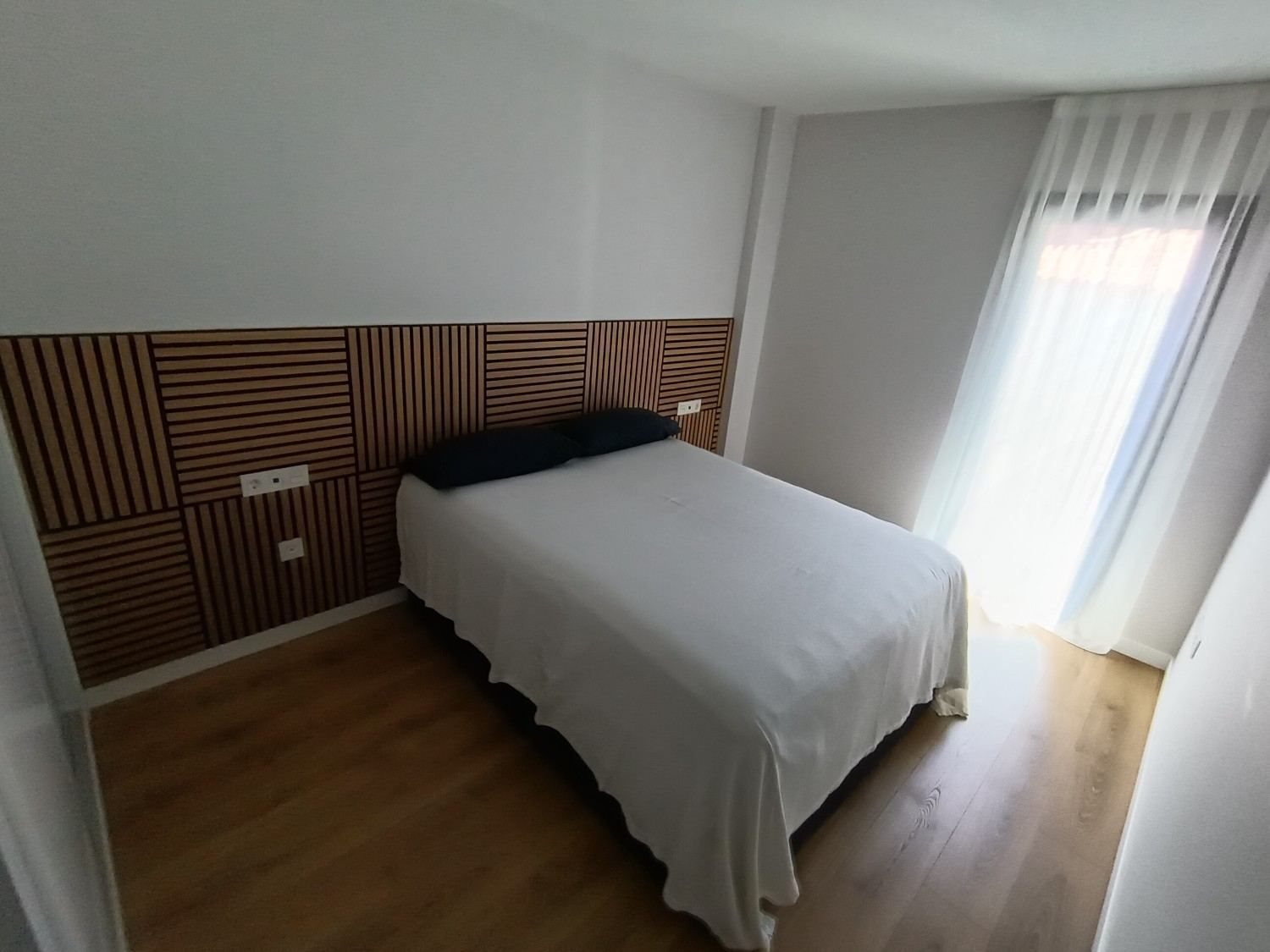 Habitación pequeña con cama, cabecero de listones de madera y luz natural que entra a través de cortinas transparentes.