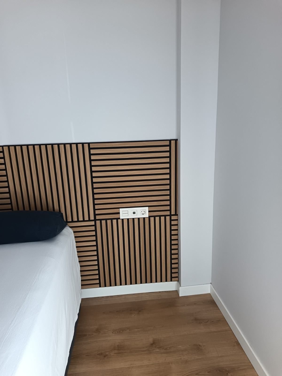 Rincón de dormitorio con cabecero de listones de madera, paredes blancas y suelo de madera.
