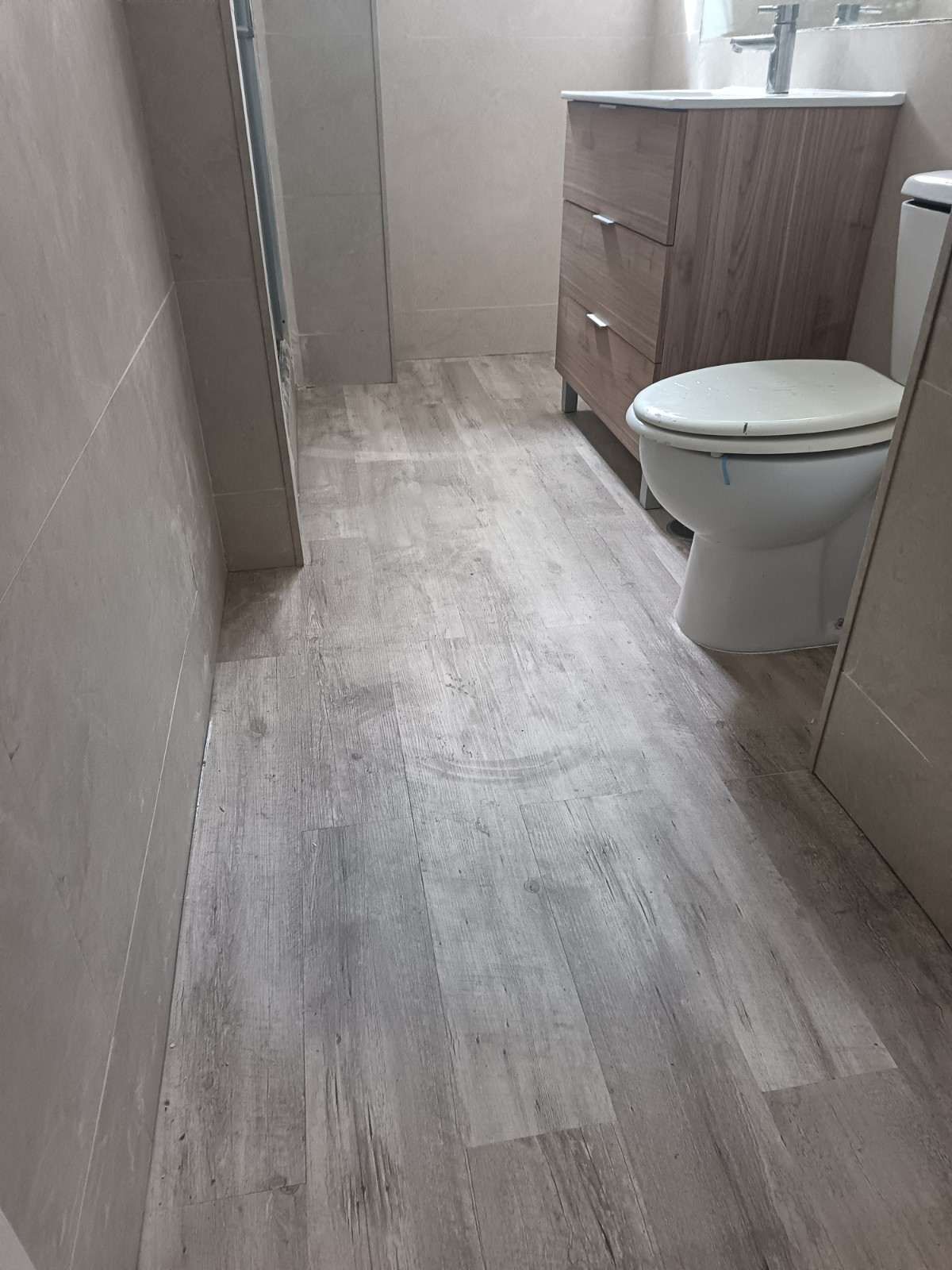 Baño estrecho con suelo de imitación madera clara, inodoro a la derecha y lavabo de madera al final.