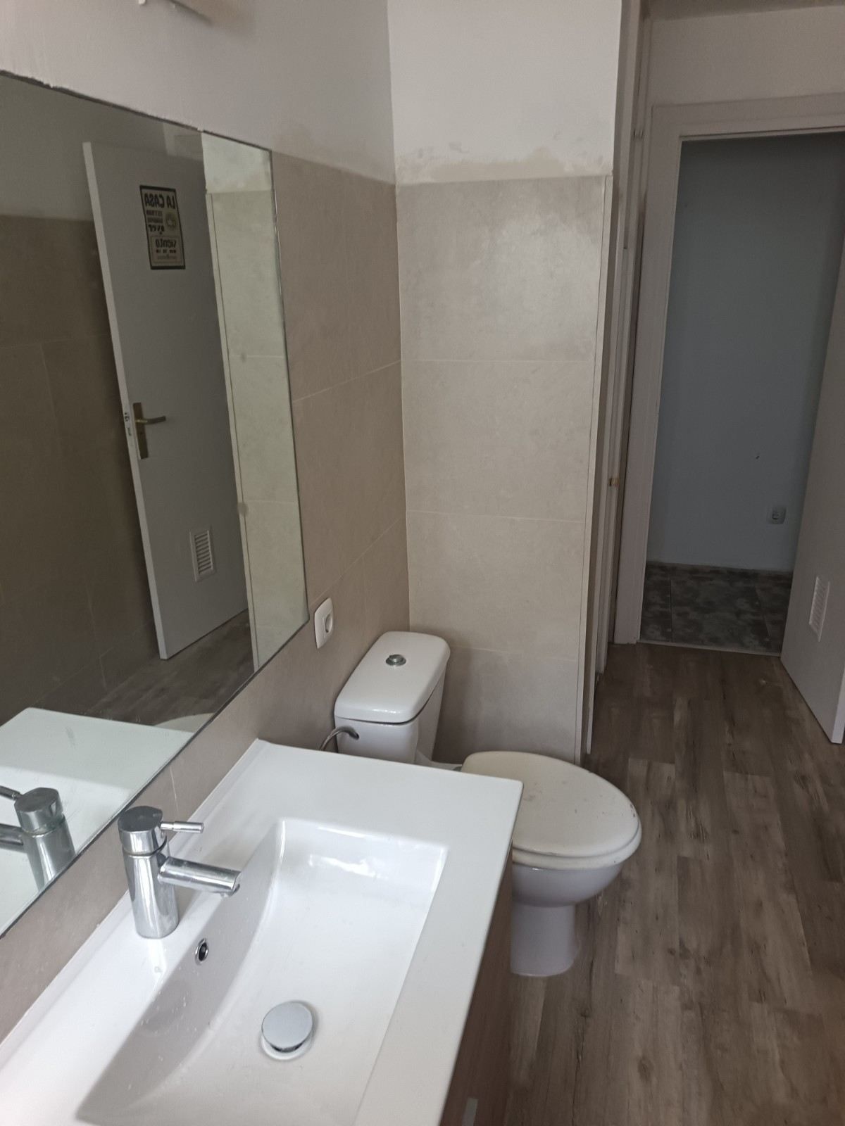 Baño pequeño con lavabo, inodoro, espejo y puertas abiertas que dan a un pasillo.