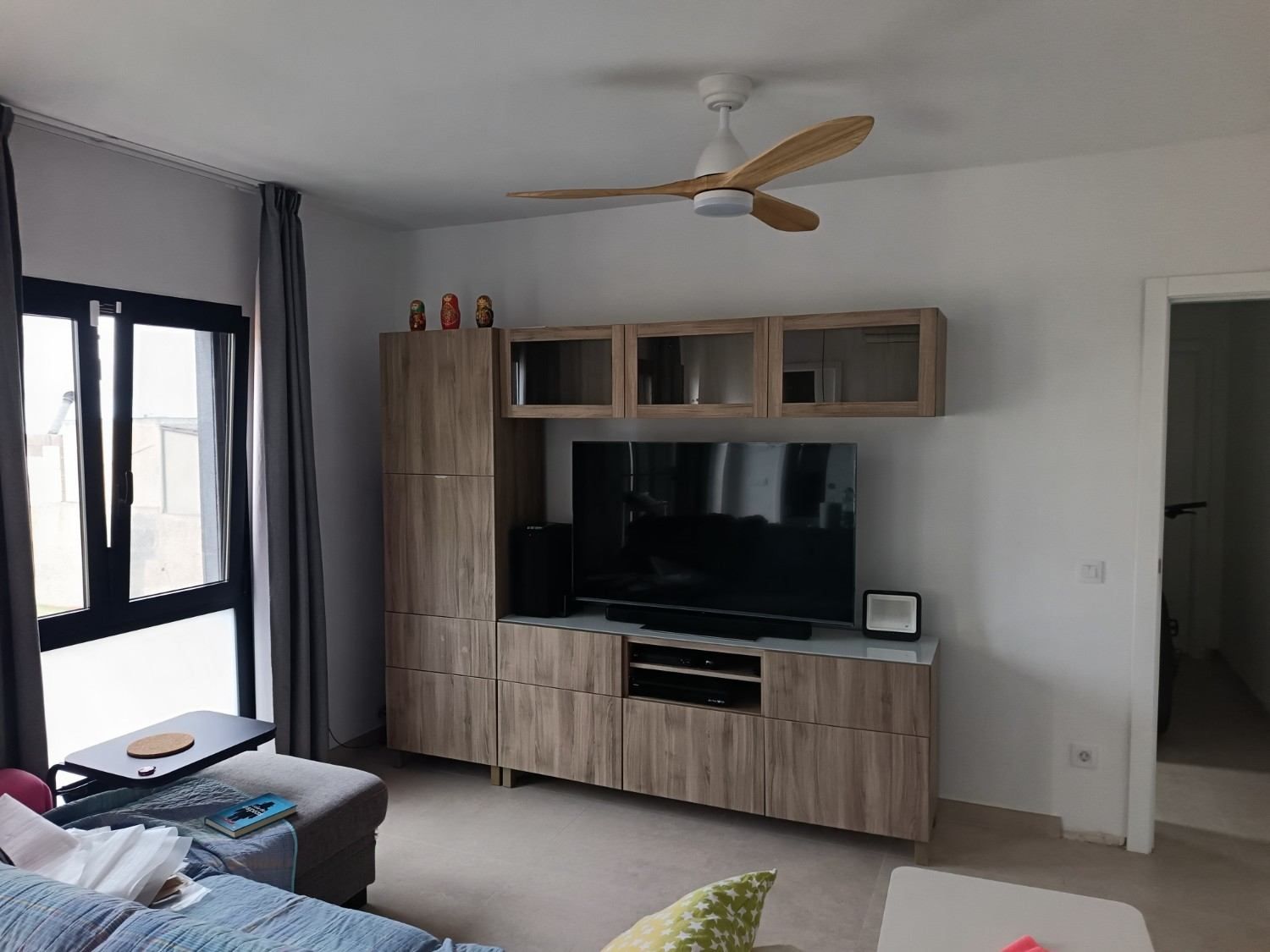 Luminoso salón con mueble para la televisión, sofá junto a grandes ventanales y ventilador de techo en una pared blanca.
