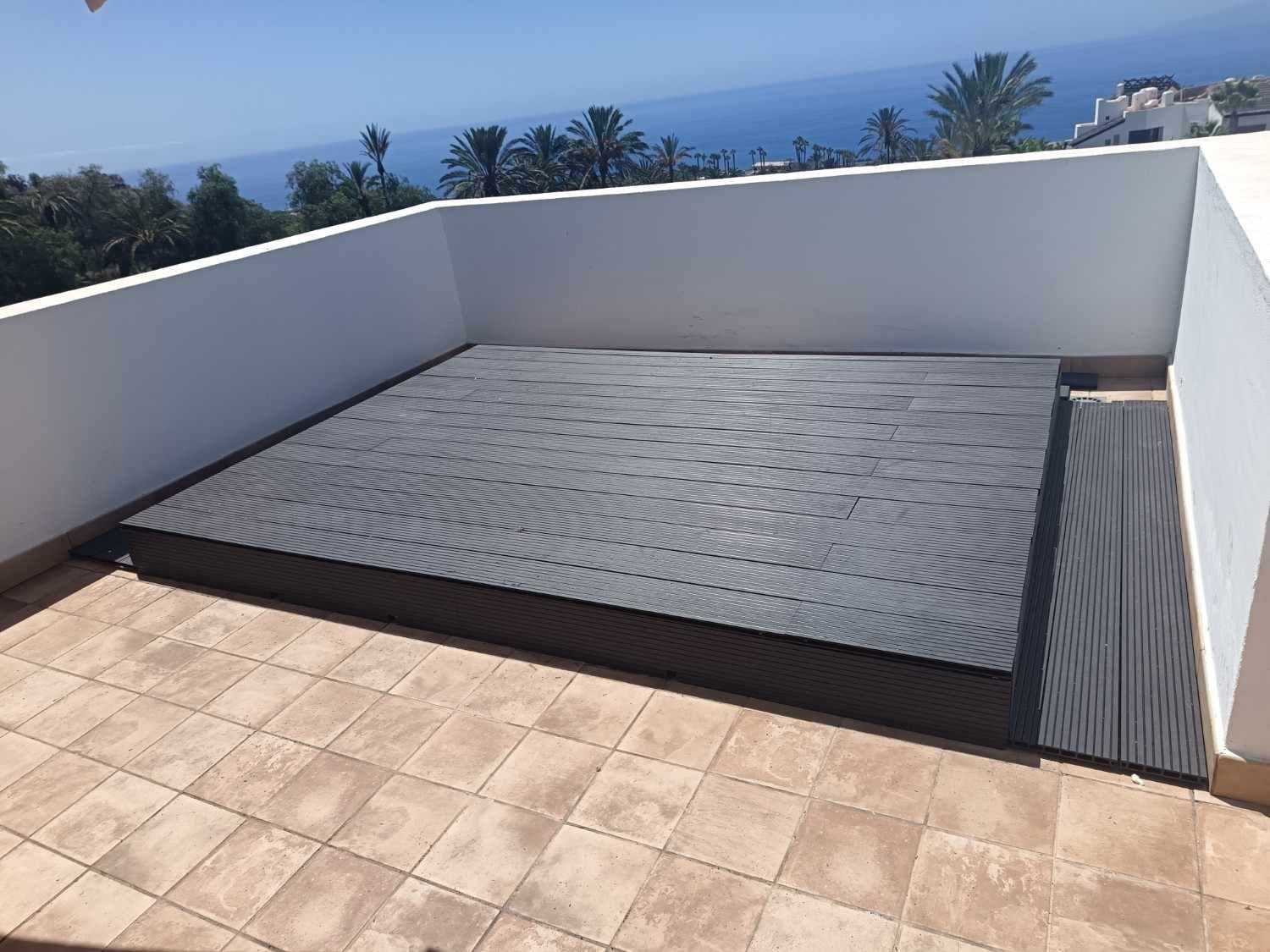 Patio exterior con un muro bajo blanco, suelo de baldosas y una tumbona cubierta con vistas al mar.