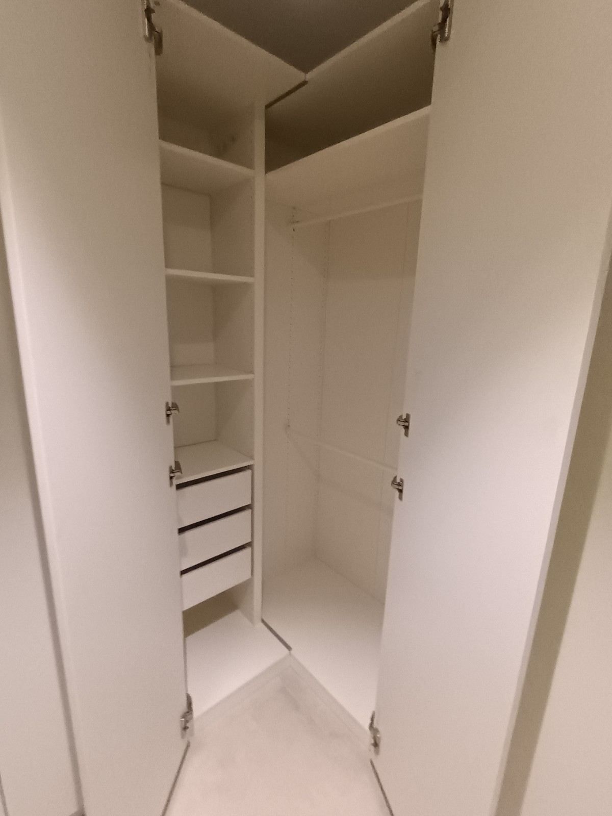 Armario esquinero blanco abierto con estantes y cajones en una habitación pequeña y vacía.
