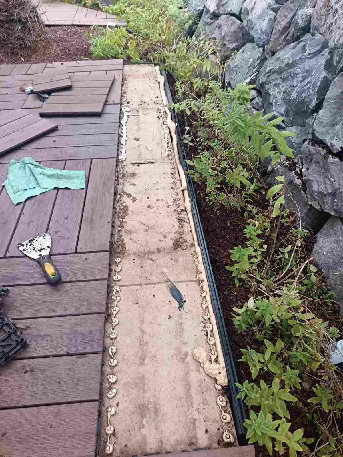 Borde de la azotea con canaleta beige, tejas oscuras, herramientas y plantas junto a un muro de piedra.