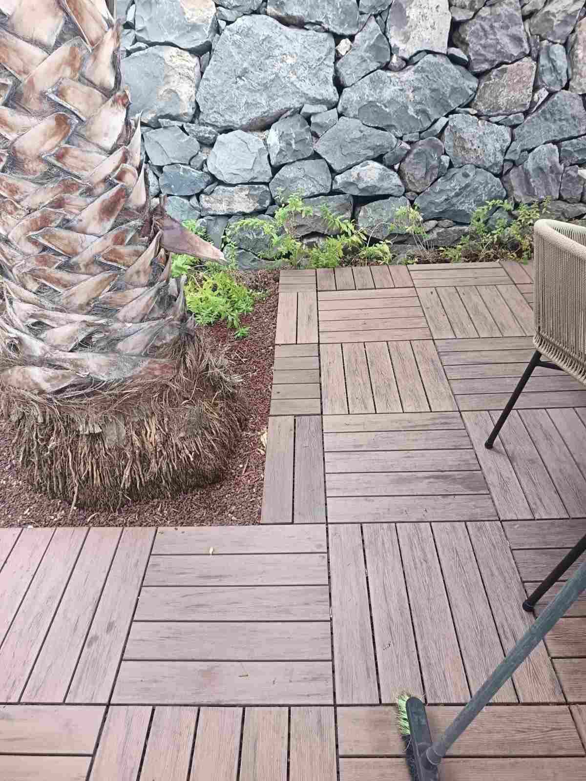 Patio con baldosas de madera, tronco de palmera, muro de piedra y una silla en la esquina.