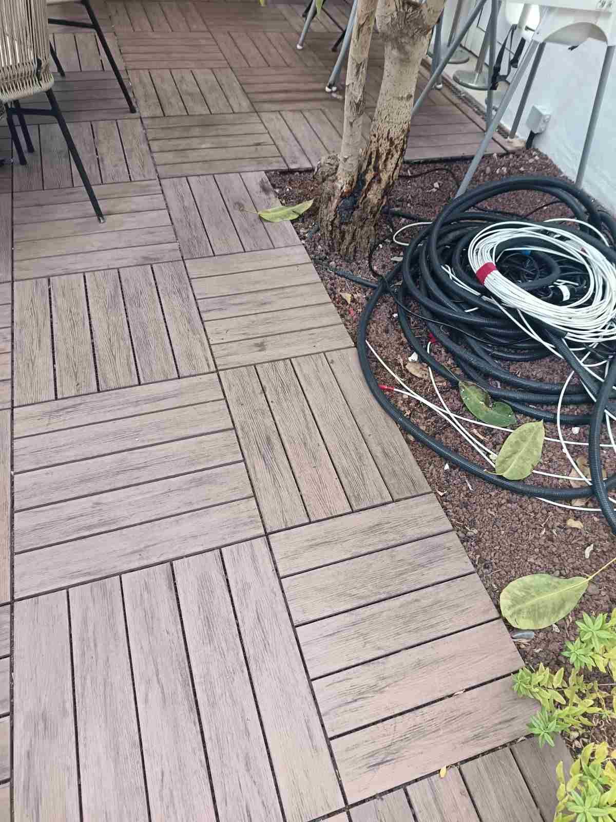 Pasarela de madera junto a un árbol, con mangueras negras enrolladas y un pequeño macizo de plantas.