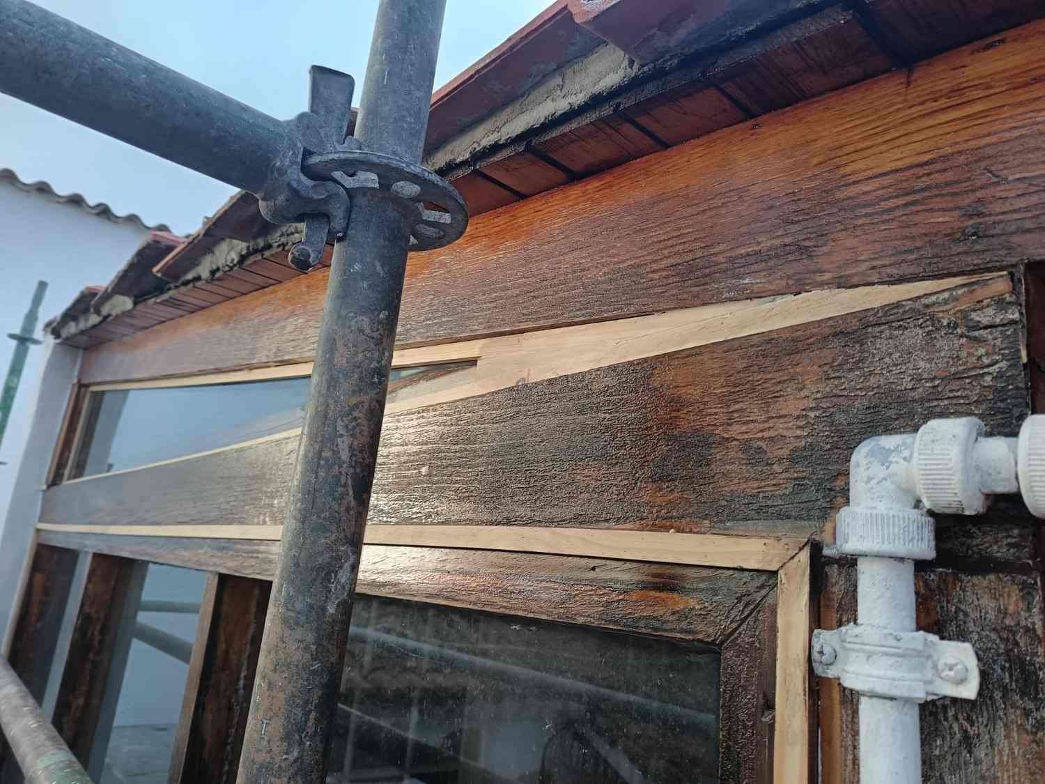 Andamios junto a una casa de madera desgastada con techo de chapa ondulada y bajantes blancos.