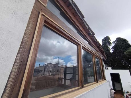 Gran ventanal con marco de madera que refleja el cielo nublado y los edificios cercanos en una terraza en la azotea.
