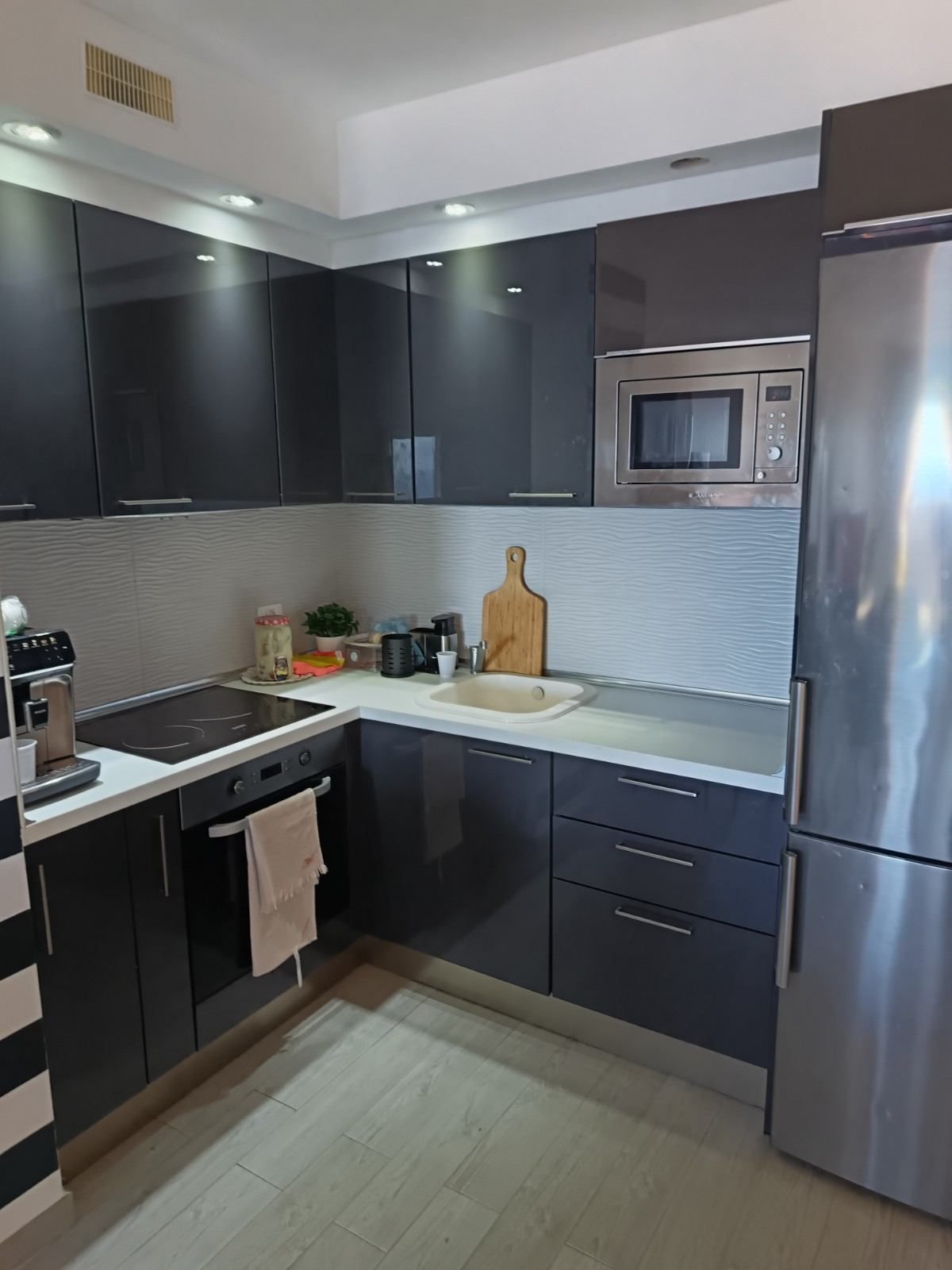 Cocina moderna con armarios oscuros, electrodomésticos de acero inoxidable, encimeras blancas y suelo de madera clara.