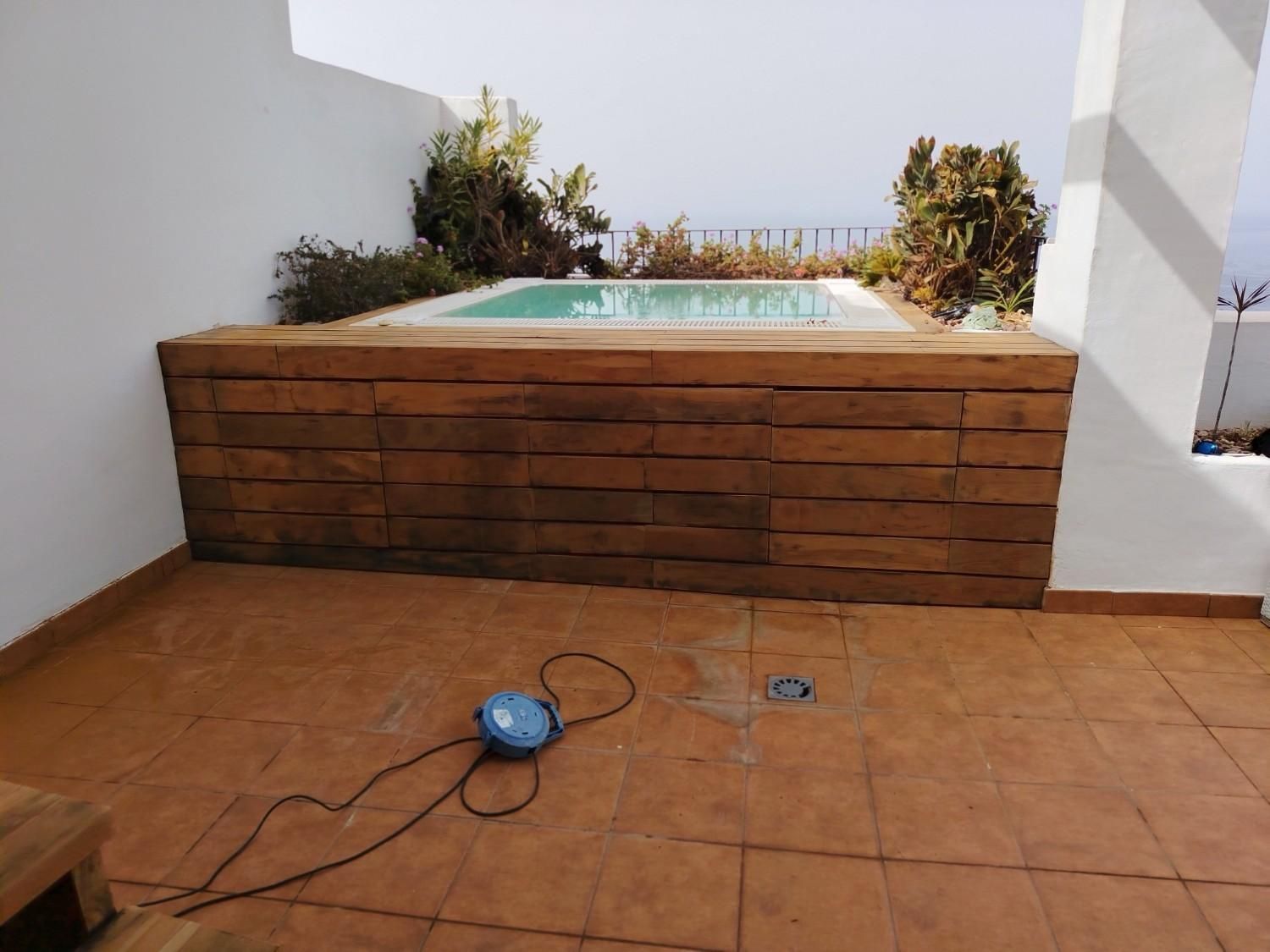 Pequeña bañera de hidromasaje en la azotea con base de madera sobre una terraza embaldosada, con una fregona azul y una manguera en el suelo.