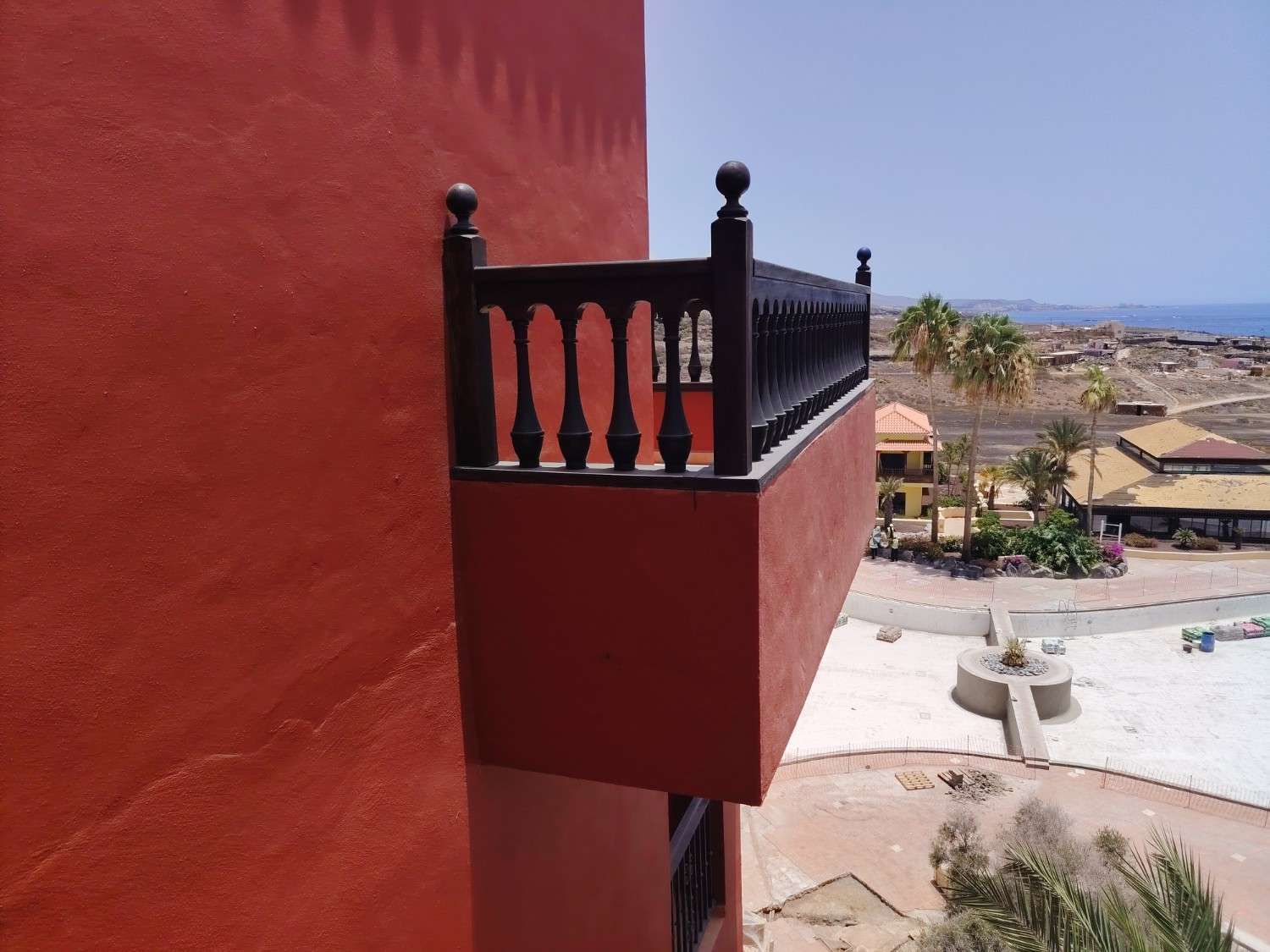 Barandilla roja en un balcón sobre una pared de terracota con vistas a una piscina y a un pueblo desértico.