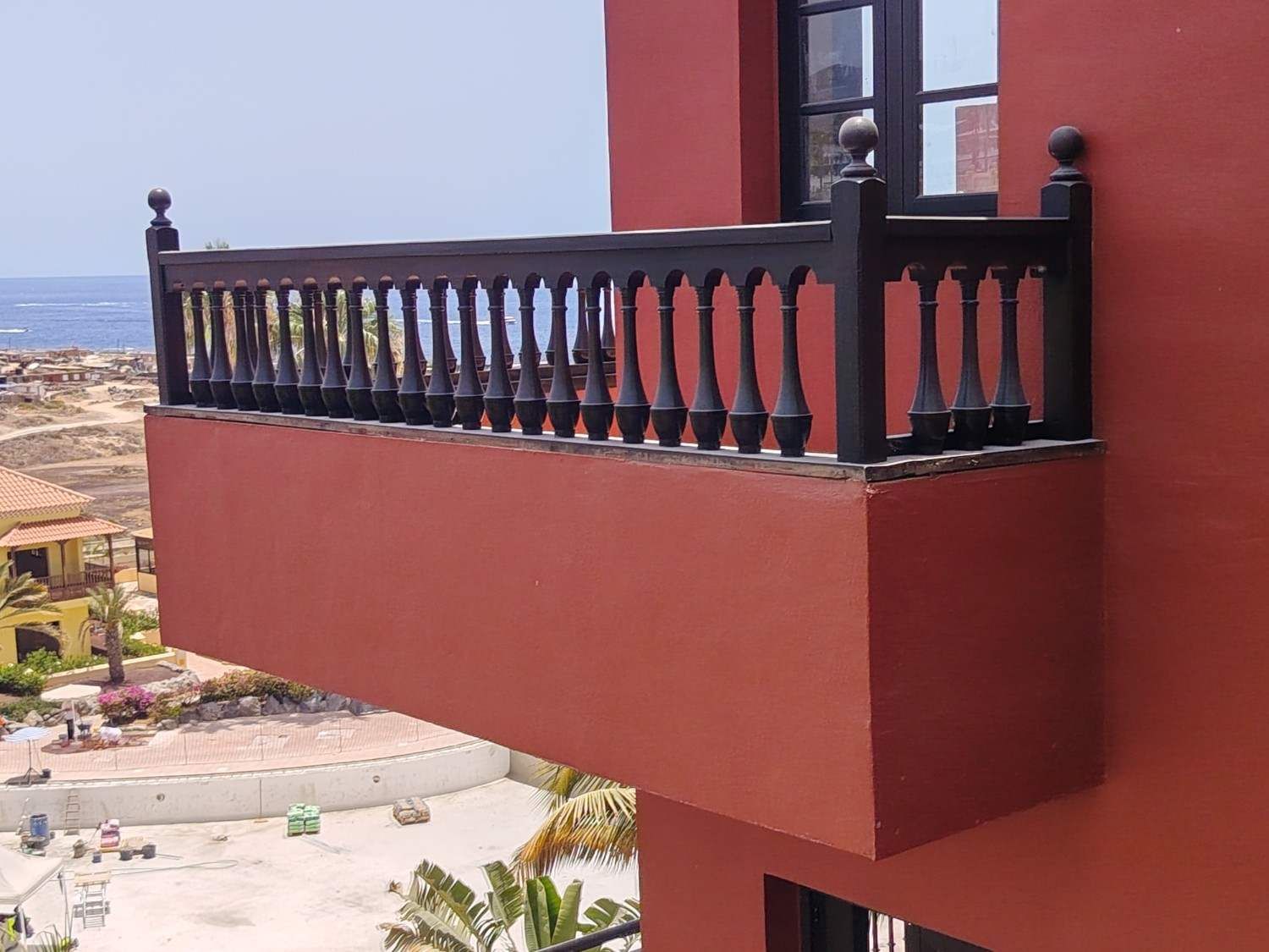 Balcón rojo con barandillas negras en un edificio con vistas a un pueblo costero y al océano.