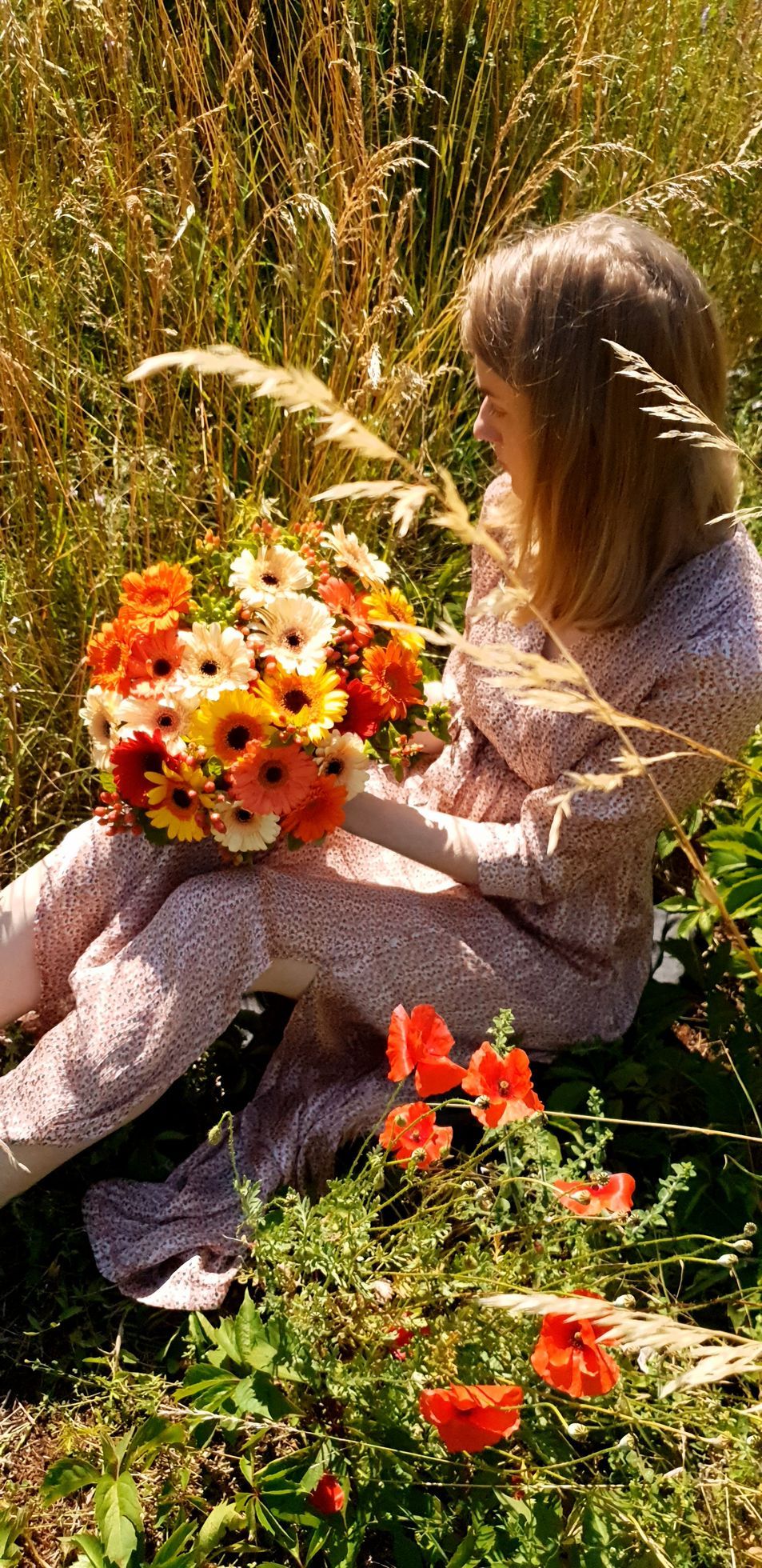 Une personne en robe blanche assise dans de hautes herbes, tenant un bouquet orange et blanc éclatant parmi les fleurs.