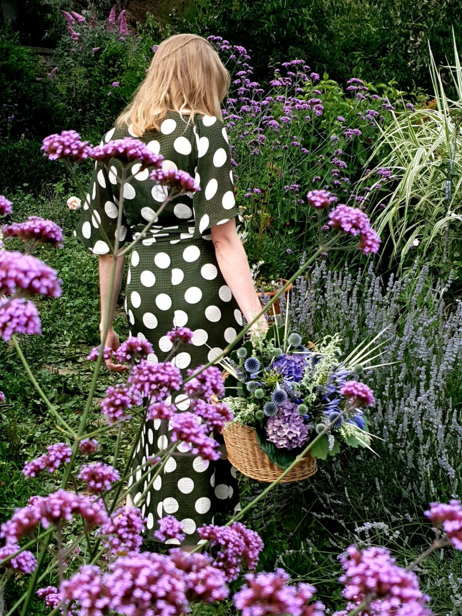 Une femme en robe verte à pois se promène dans un jardin luxuriant orné de fleurs violettes et portant un panier.