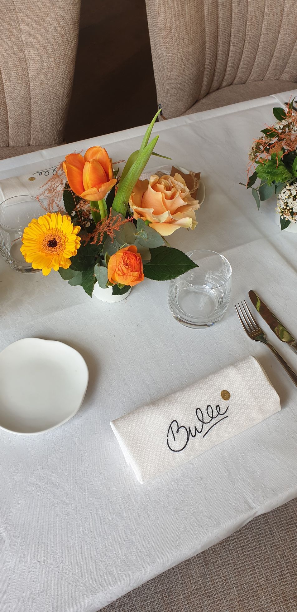 Composition florale et couvert sur une nappe blanche avec une carte nominative manuscrite.