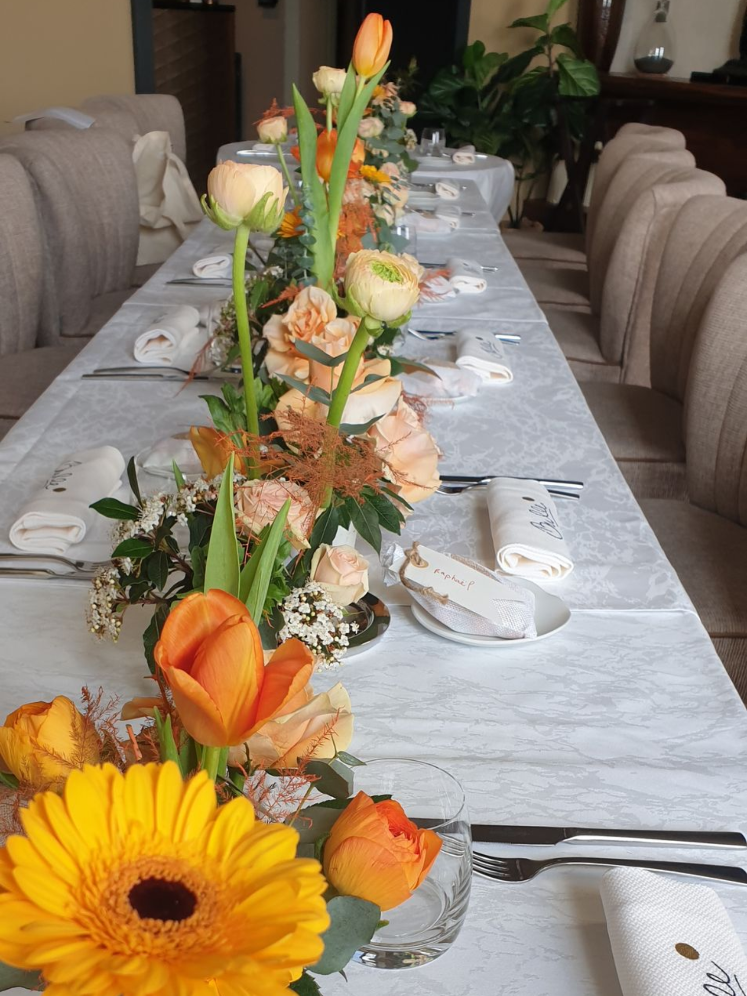 Longue table de banquet avec nappes blanches et centres de table floraux orange, dressée avec couverts et chaises.