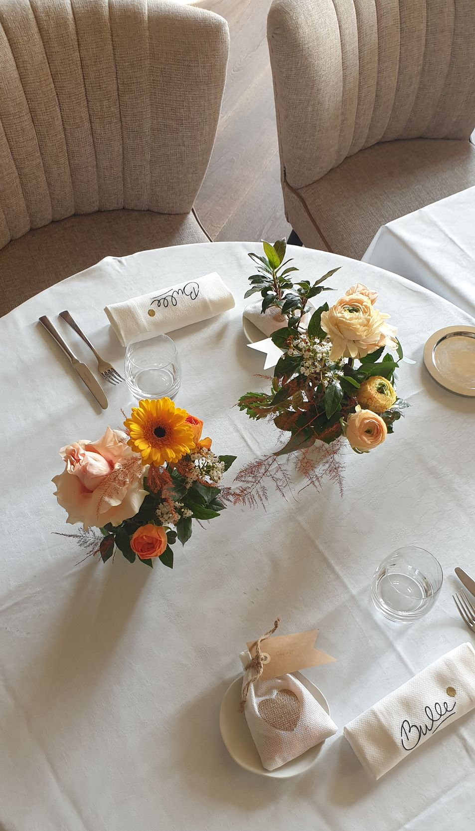 Table ronde ornée de petits centres de table floraux, de couverts et de serviettes pliées, dans une salle à manger lumineuse.