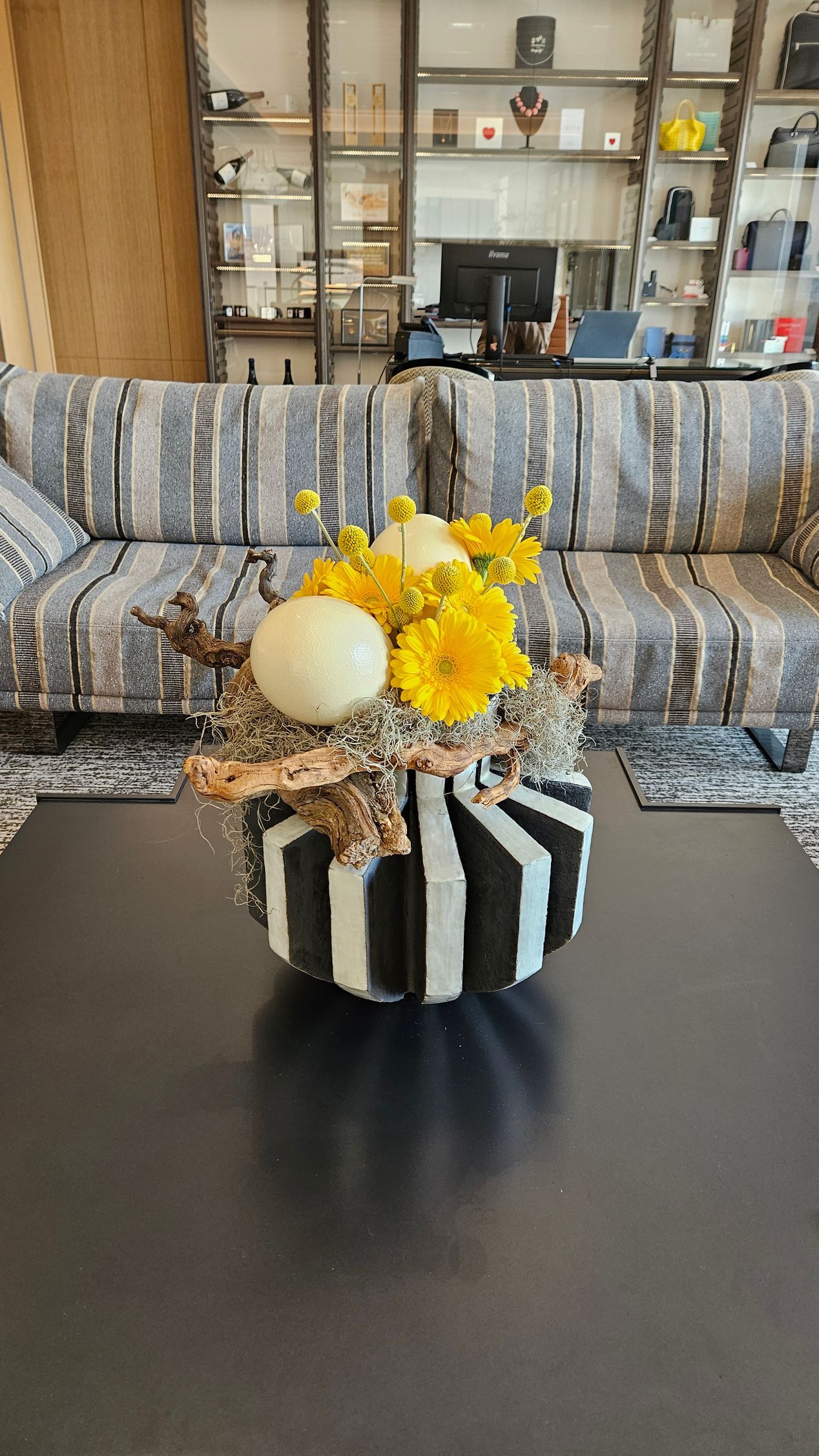 Des fleurs jaunes dans un vase rayé sur une table noire, avec un canapé et une vitrine en arrière-plan.