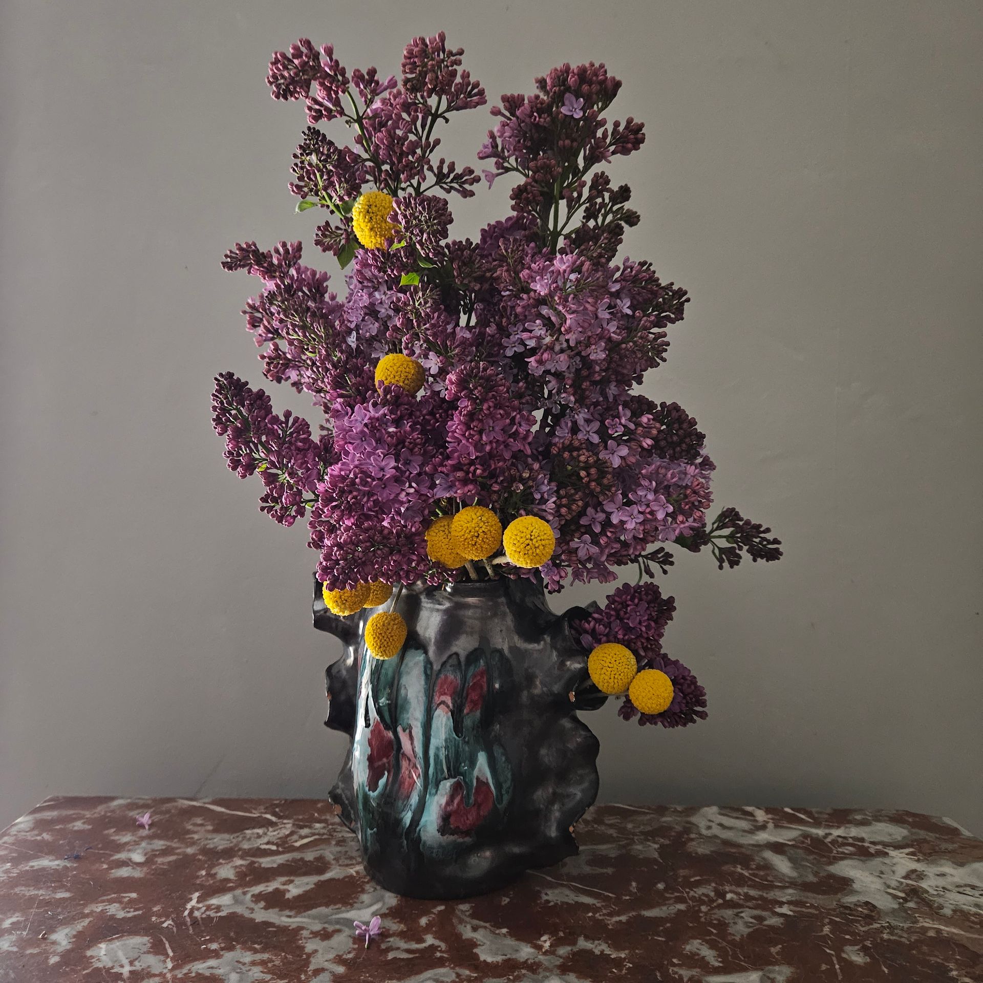 Des fleurs violettes et des fleurs jaunes dans un vase décoratif sombre, posé sur une table devant un mur uni.