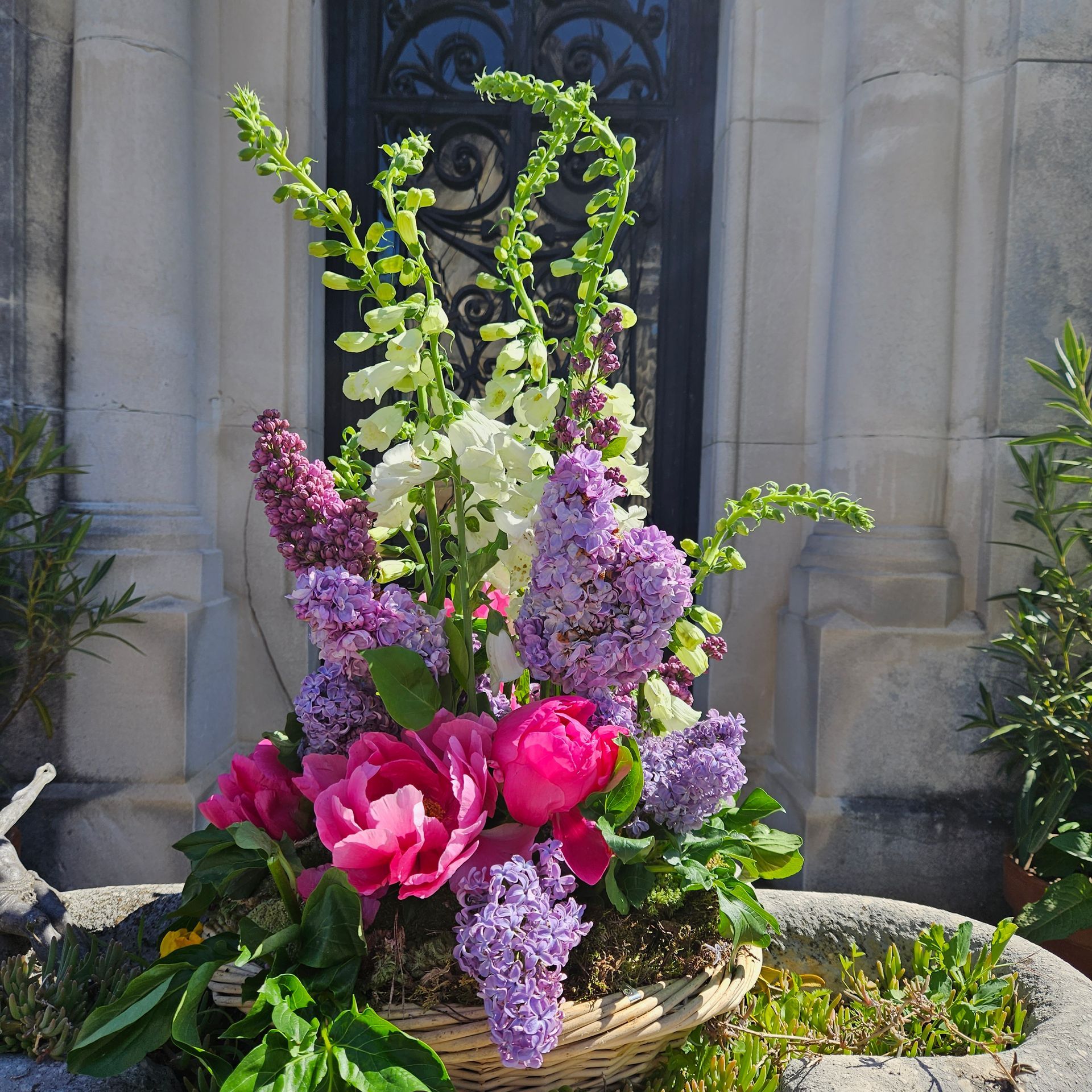 Composition florale colorée avec des roses roses, des fleurs violettes et de la verdure dans un panier devant un bâtiment en pierre