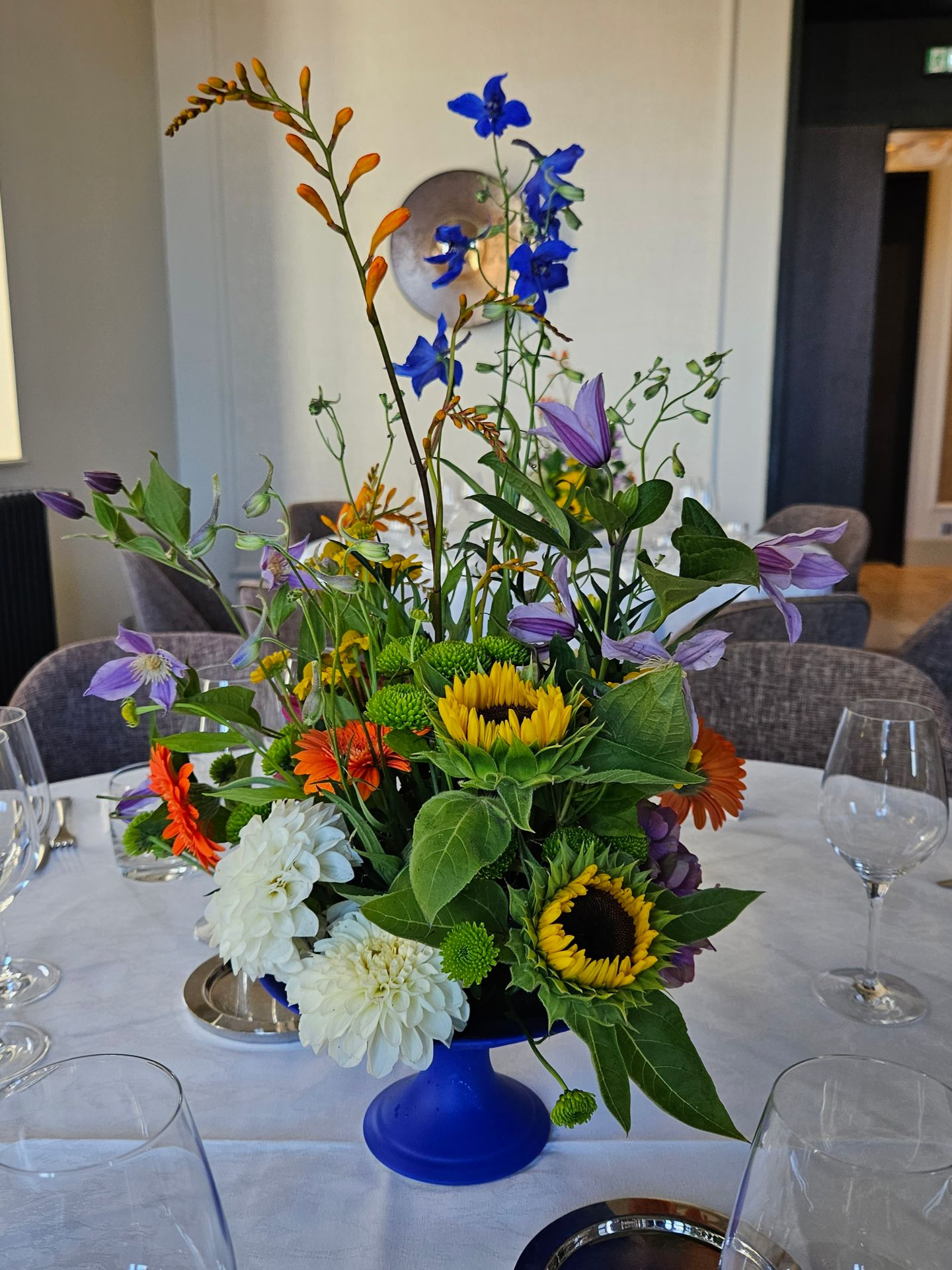 Un centre de table floral coloré dans un vase bleu, avec des tournesols et des marguerites blanches, orne une table à manger dressée.