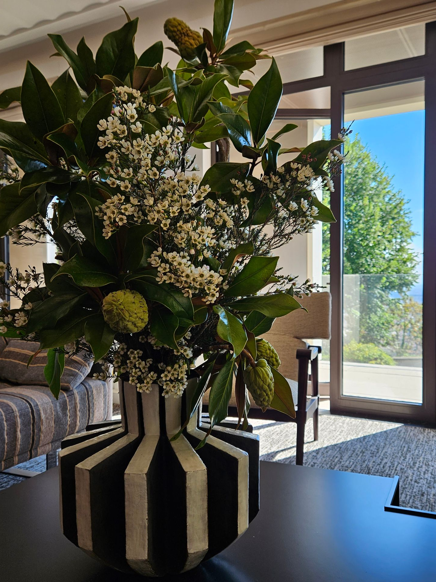 Grand arrangement floral dans un vase à rayures noires et blanches sur une table près d'une fenêtre lumineuse