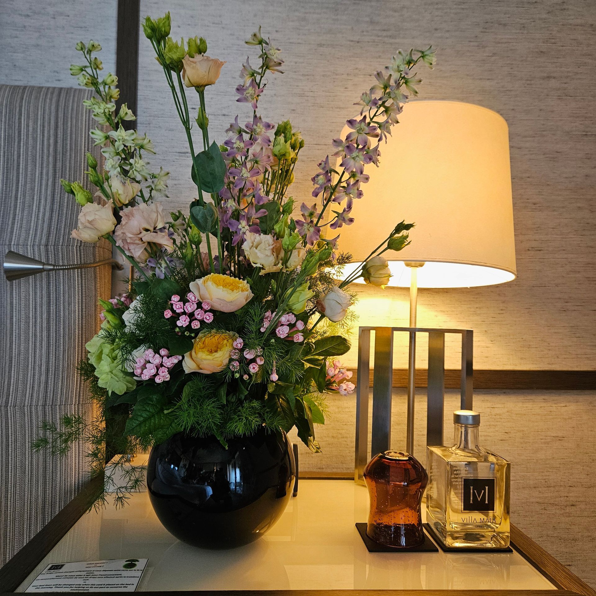 Composition florale dans un vase noir, à côté d'une lampe de table allumée et de bougies sur une console.