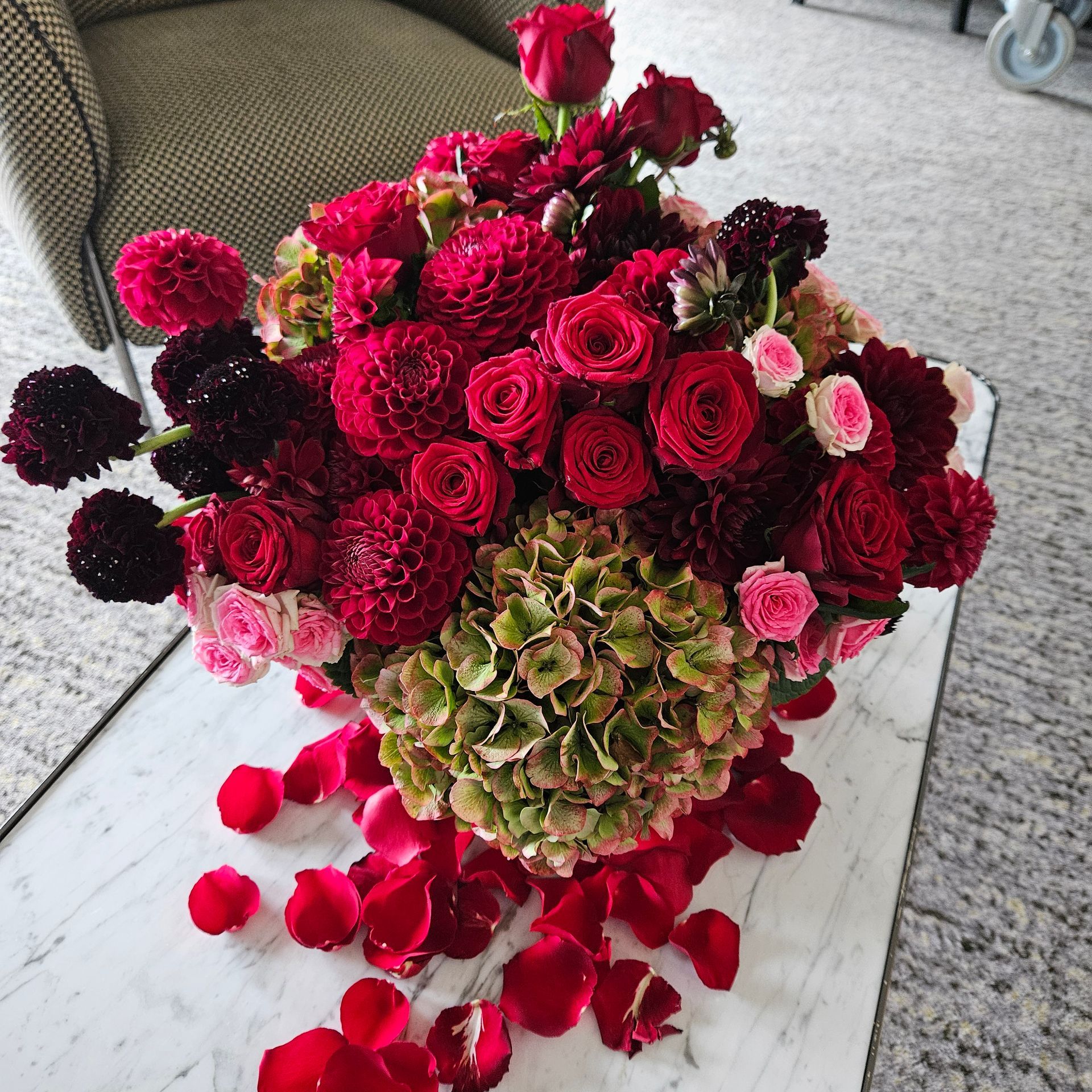 Bouquet de fleurs rouges, roses et bordeaux, avec des pétales de rose débordant sur une table blanche.