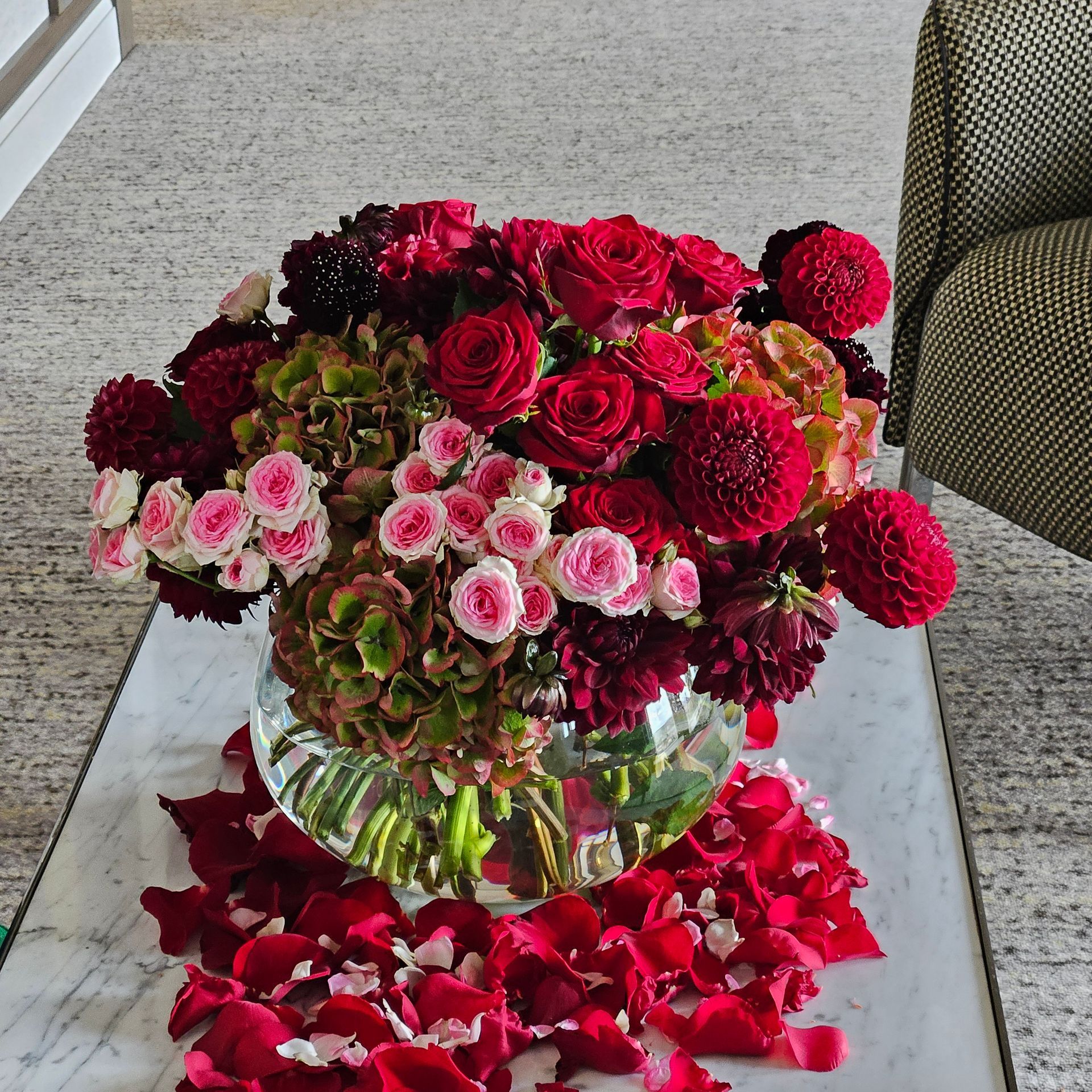 Bouquet de fleurs rouges et roses mélangées dans un vase en verre, posé sur un sol recouvert de moquette, avec des pétales rouges éparpillés.