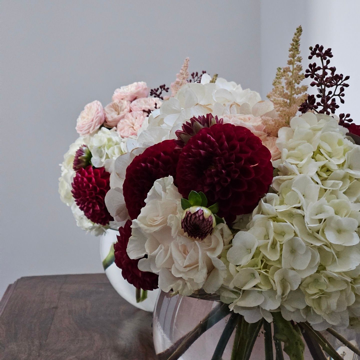 Vase blanc contenant des fleurs bordeaux, rose poudré et crème posé sur une table en bois devant un mur clair.