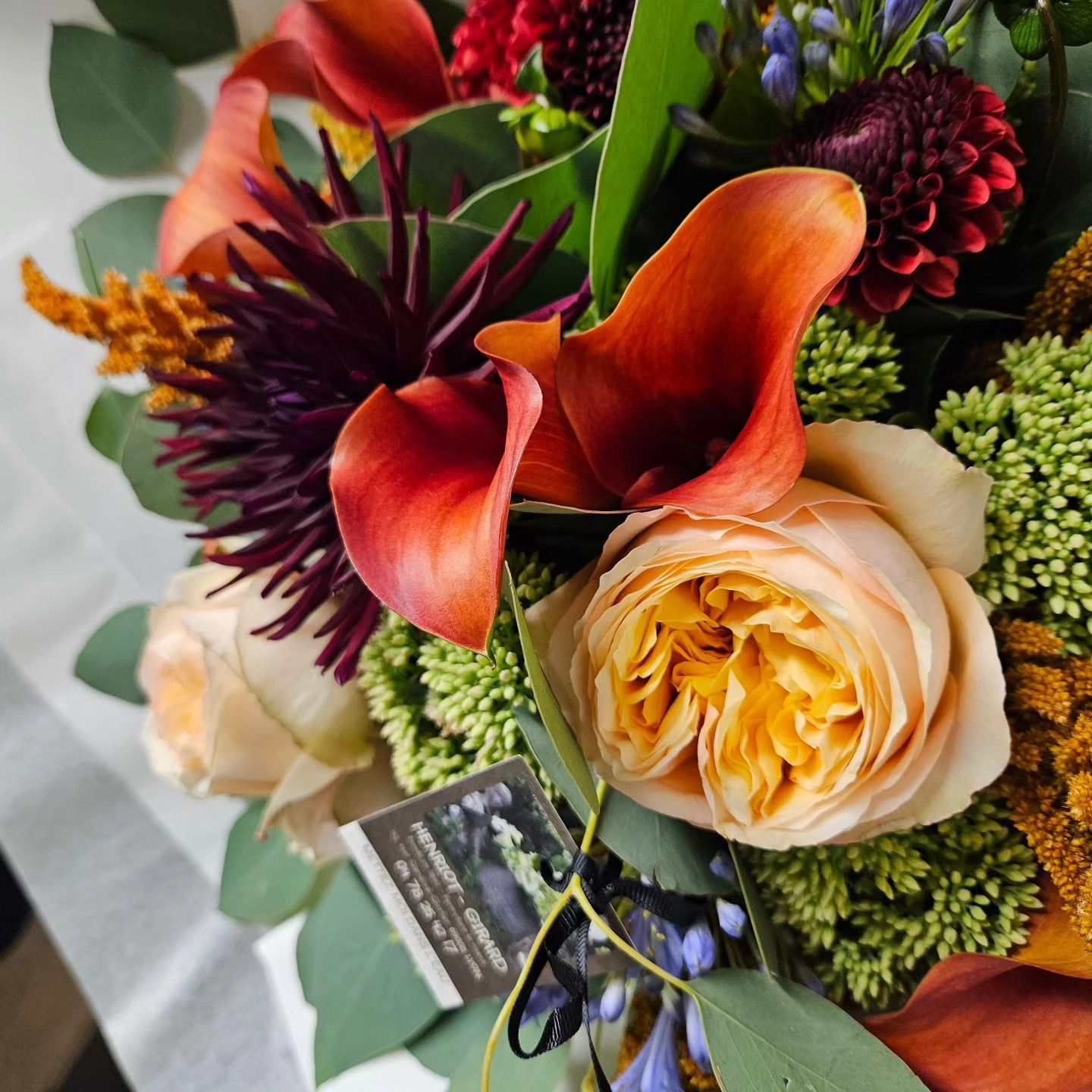Bouquet coloré composé de callas orange, de roses crème et de fleurs piquantes violet foncé